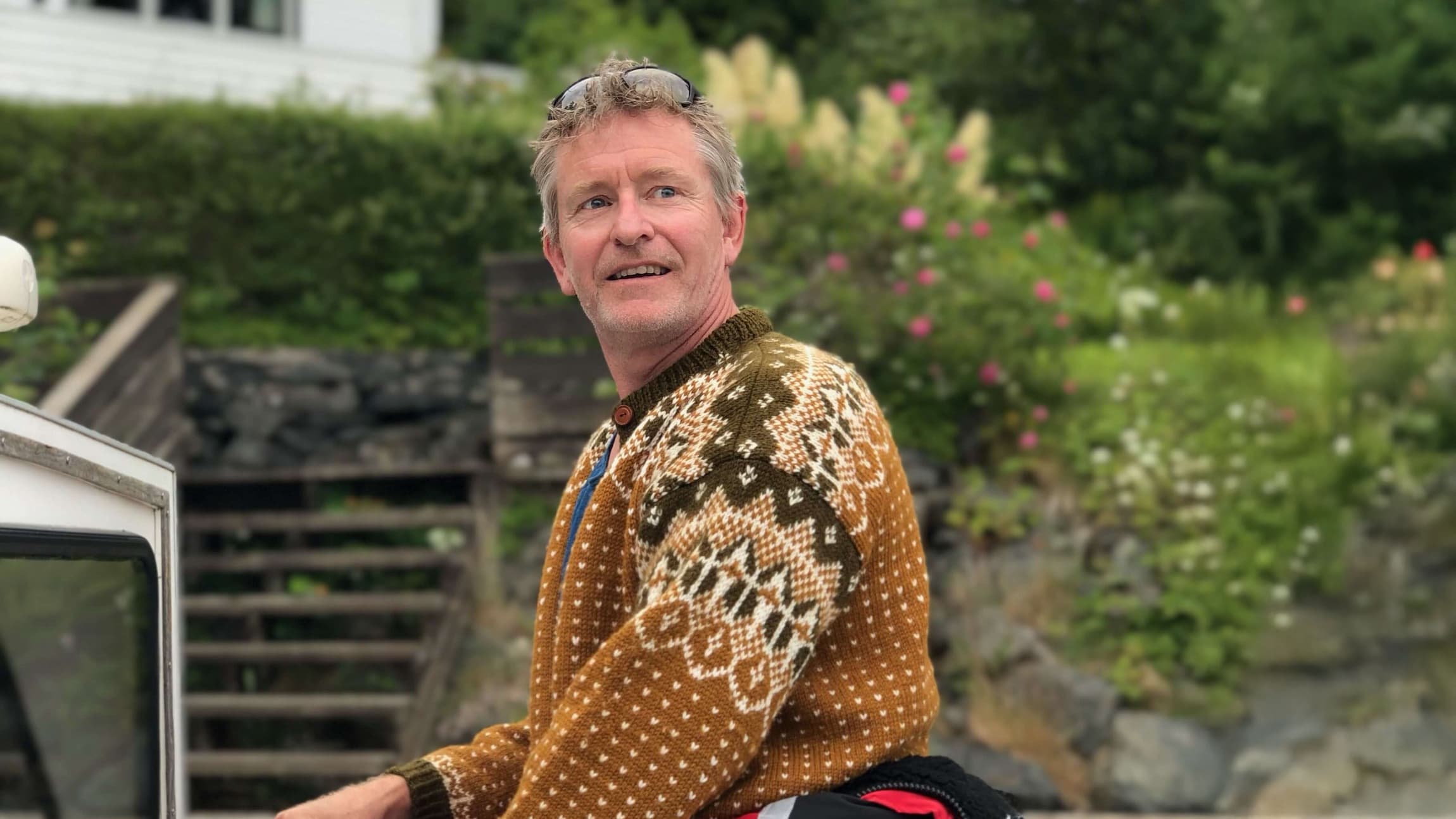 Skuespiller Henrik Mestad står til rors i en båt. Han har på seg brun strikkegenser og en rød kjeledress som er knyttet rundt livet.