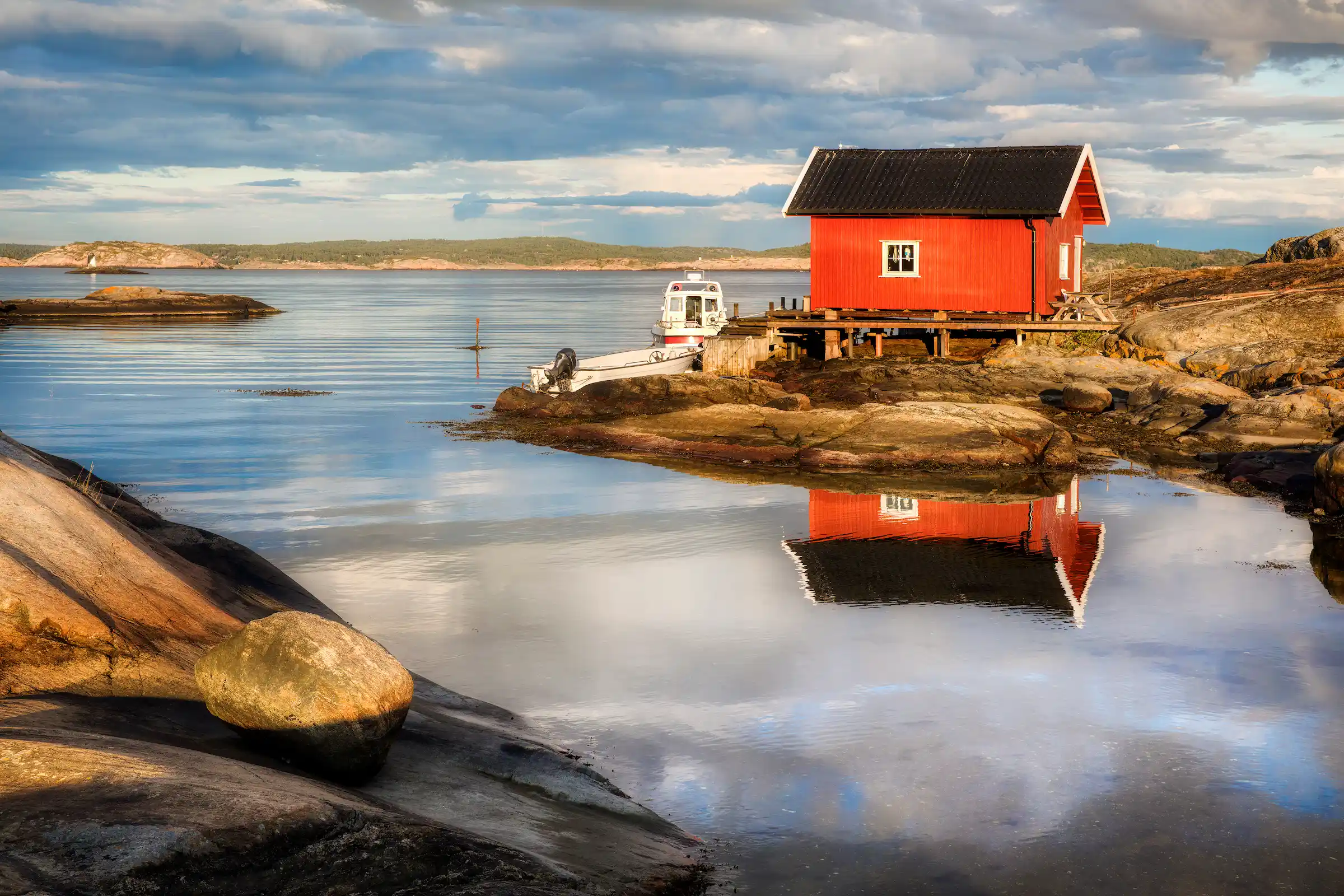 bilde fra land ut mot en stille skjærgård med en gammel trebrygge og to båter. et rødt båthus står på berget ved brygga.