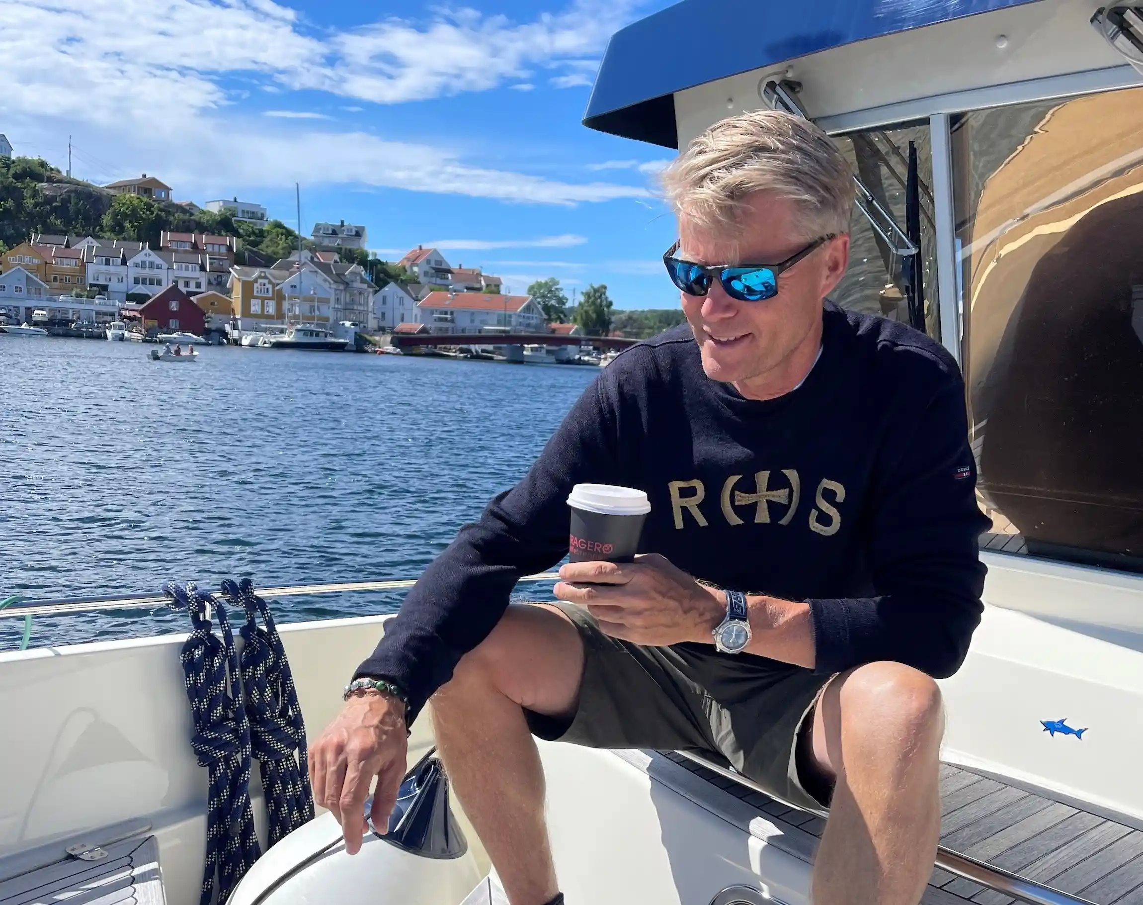 Øyvind sitter i båt med kaffe, ser ned og smiler