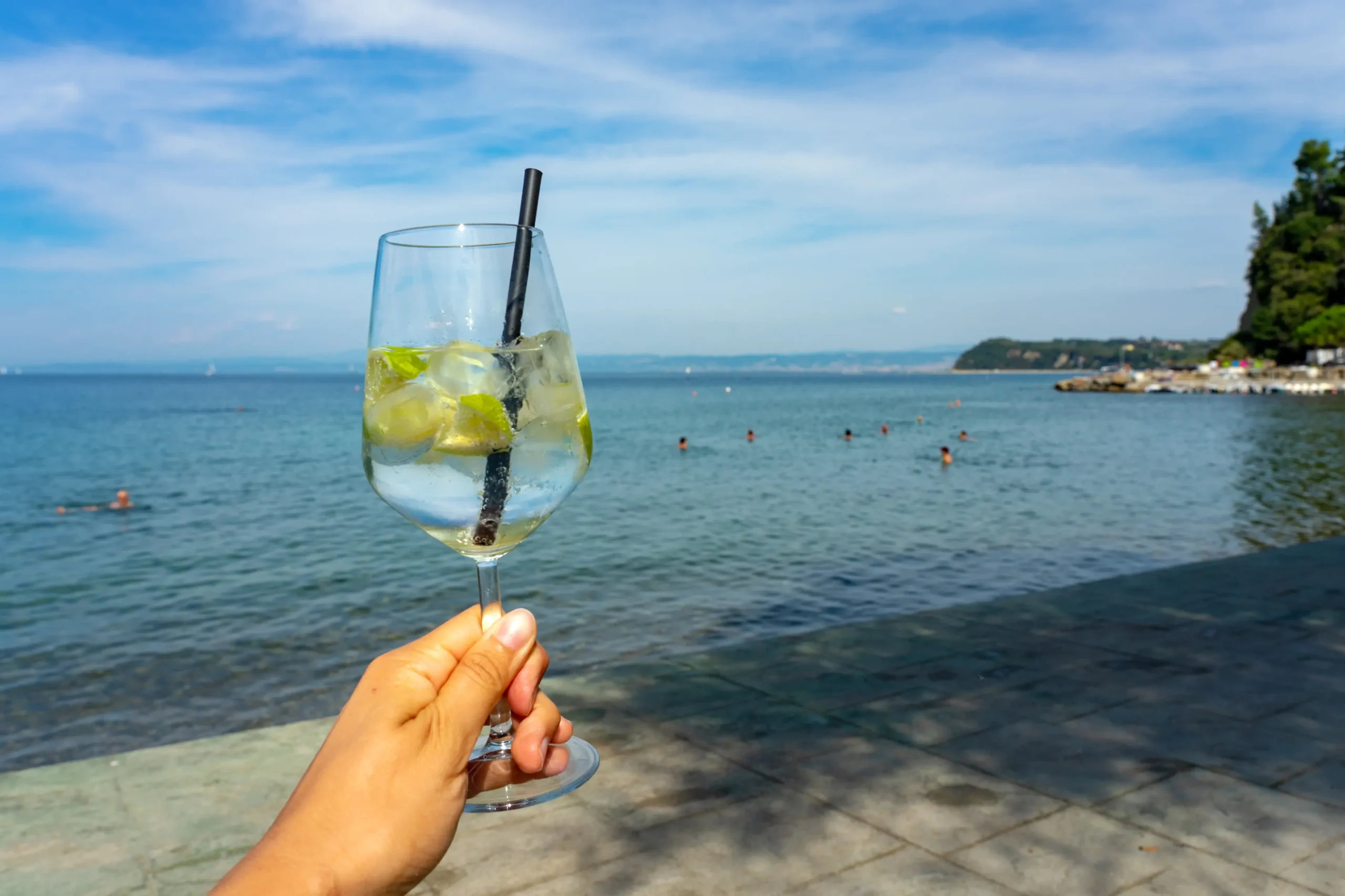 En hånd holder et glass med en limedrink. I bakgrunnen ser vi en strand med folk som bader.