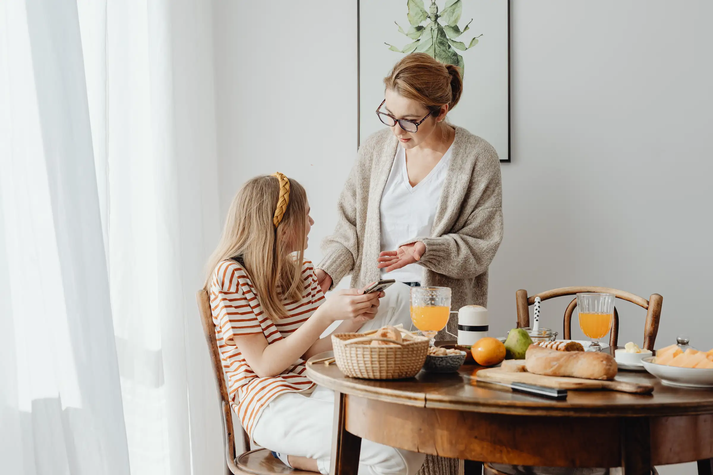 Mor og datter som sitter ved frokostbordet og snakker om hva som skjer på mobilen. Moren står over jenta og ber om å få se på mobilen.