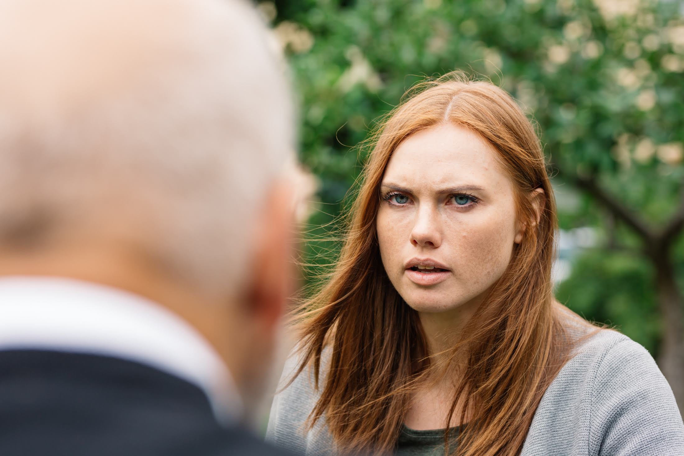 Kvinne med rødt hår og grå cardigan-genser ser bekymret mot en eldre mann, kledd i hvit skjorte med blå genser over, som vi bare ser bakhodet til. De ser ut til å ha en alvorlig prat.