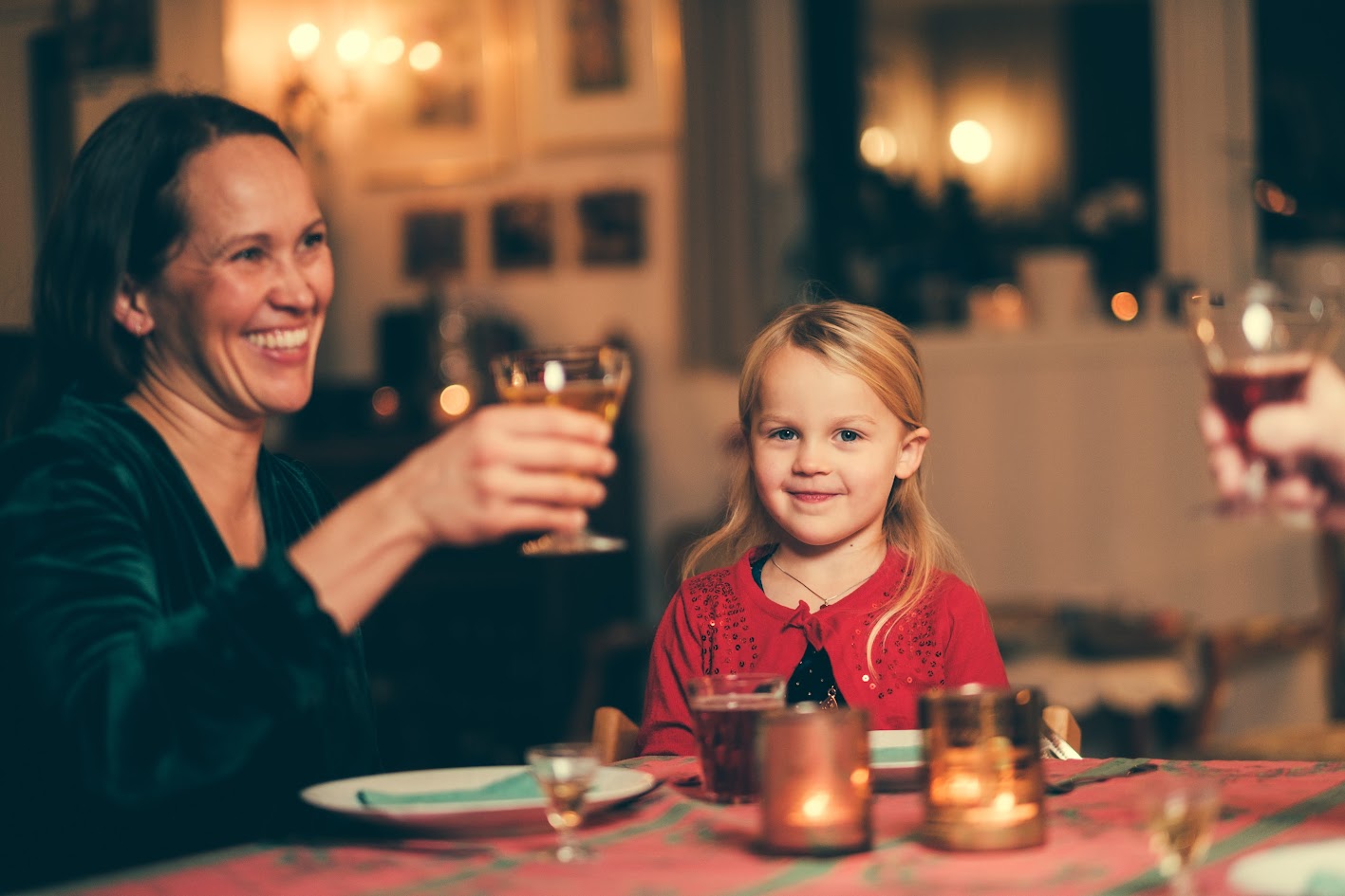 Julepyntet jente sitter ved bordet og ser smilende inn i kamera. Ved siden av henne sitter mot og smiler og skåler.