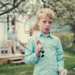 En gutt står i en sommerhage og holder to badmintonracketer. Han ser alvorlig inn i kamera.