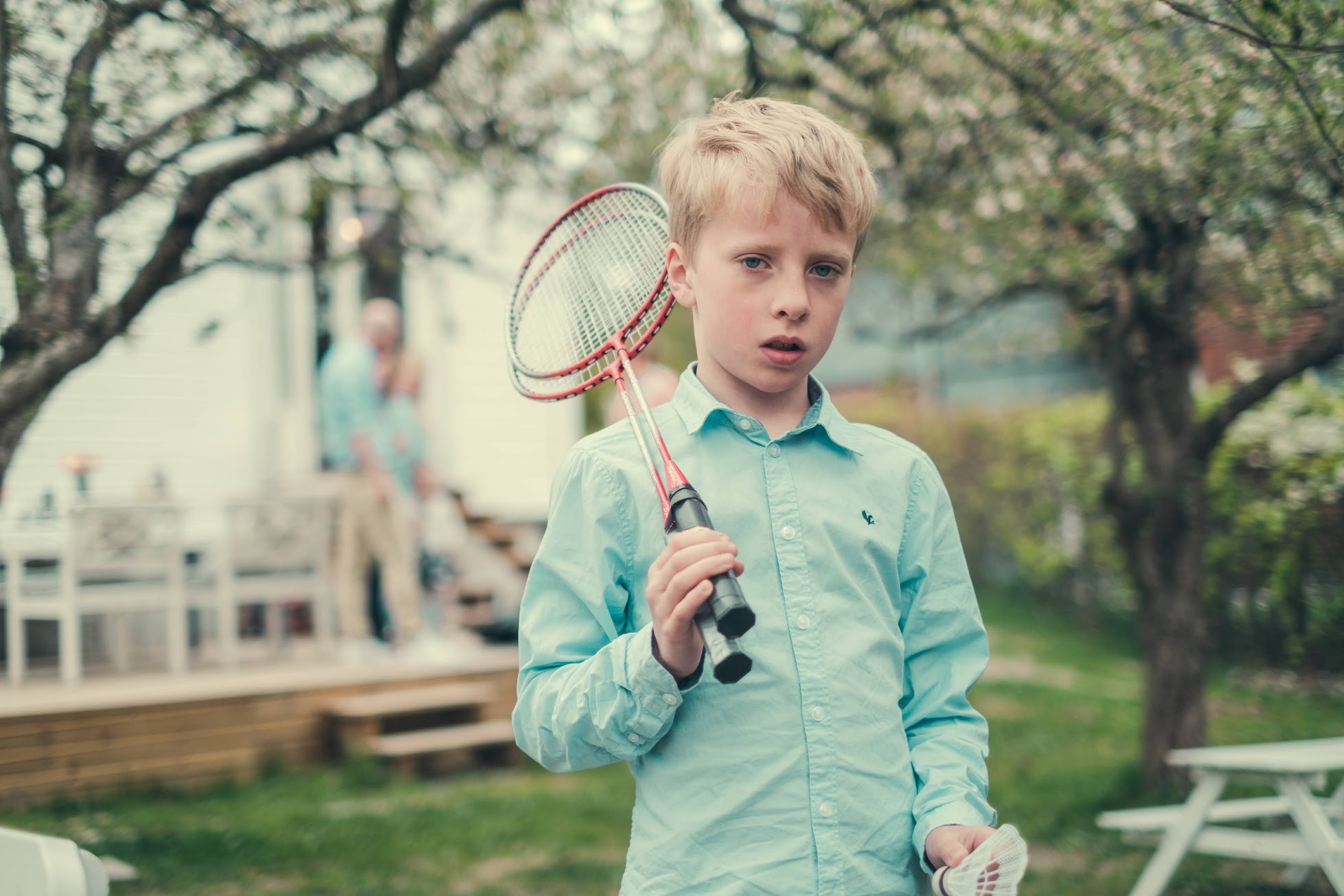 En gutt står i en sommerhage og holder to badmintonracketer. Han ser alvorlig inn i kamera.