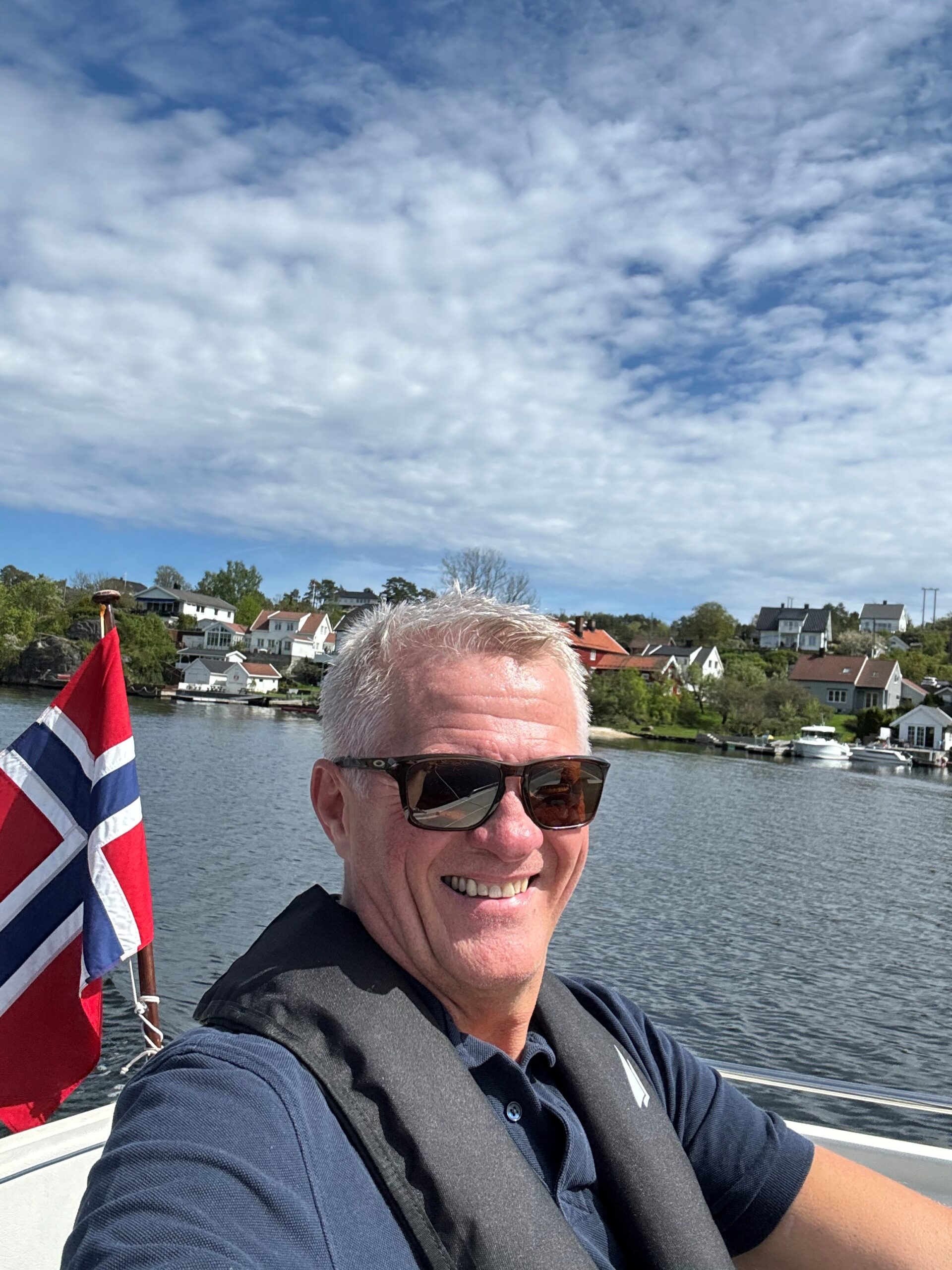 Stig Johnsen. Mann avbildet på sjøen. Har på seg solbriller og smiler. Norskt flagg i bakgrunnen