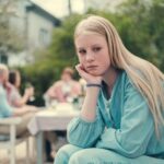 Tenåringsjente sitter ute på en terrasse og ser til siden med et ettertenksomt uttrykk. I bakgrunnen sitter det voksne folk med glass i hendene.