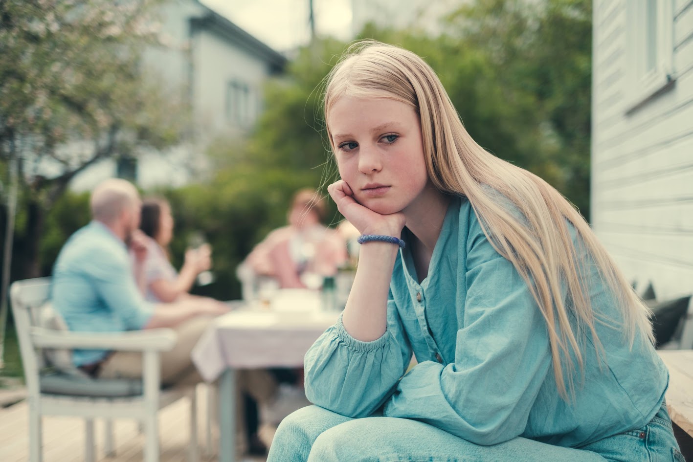 Tenåringsjente sitter ute på en terrasse og ser til siden med et ettertenksomt uttrykk. I bakgrunnen sitter det voksne folk med glass i hendene.
