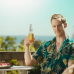 En mann i hawai-skjorte sitter utendørs i sola med en flaske i hånda. Han ser i kamera med et ironisk blikk.