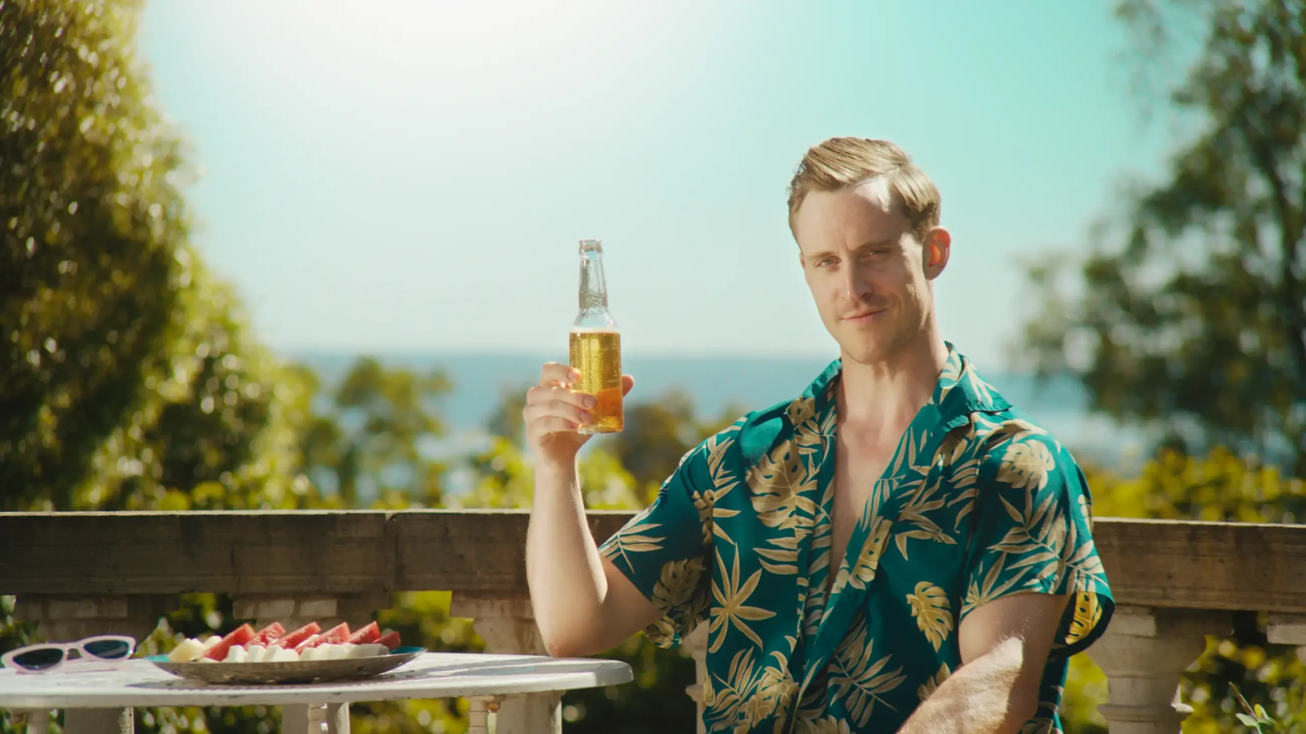 En mann i hawai-skjorte sitter utendørs i sola med en flaske i hånda. Han ser i kamera med et ironisk blikk.