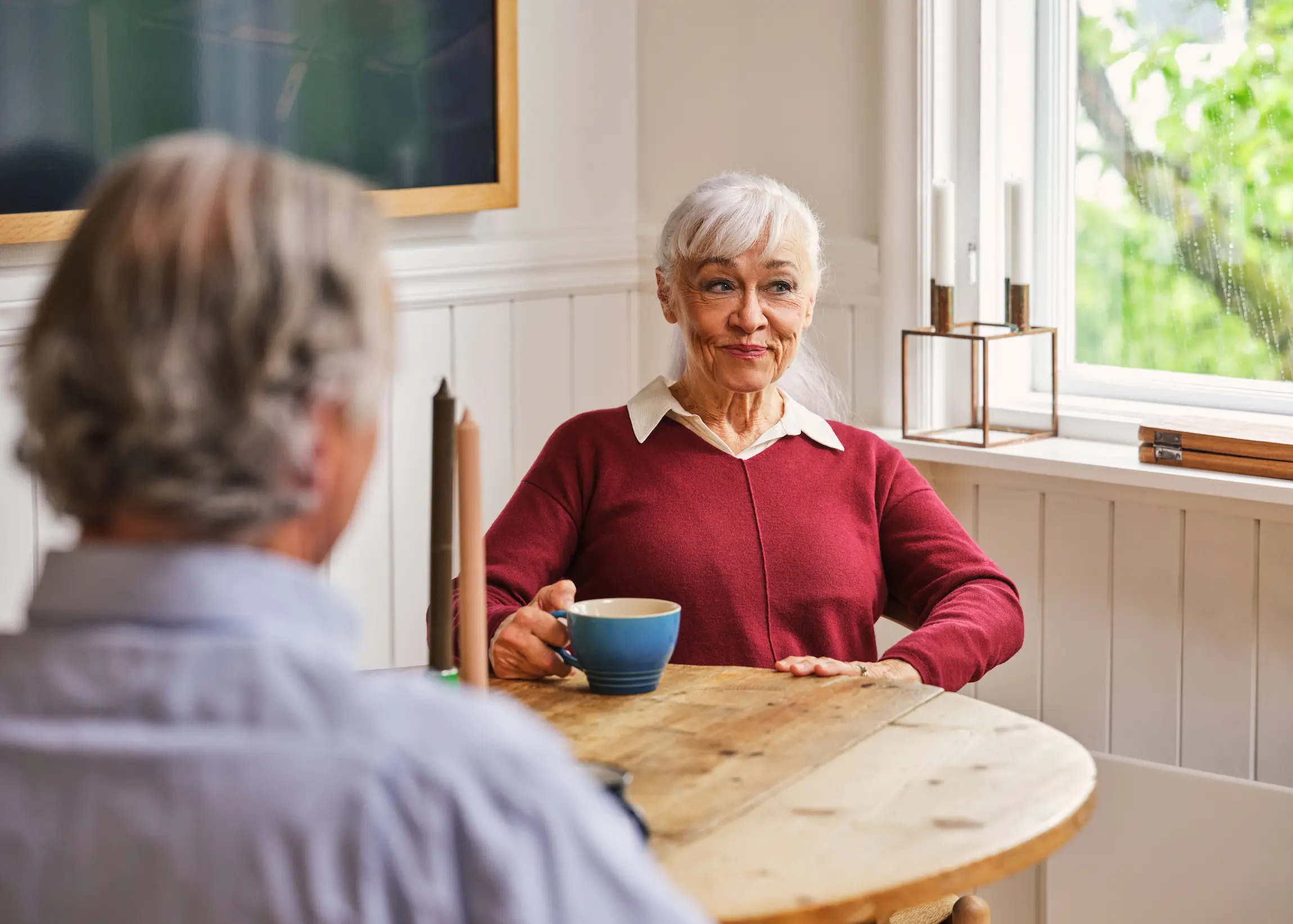 Kvinnelig senior sitter rundt et bord med rød genser og en stor kopp. I forgrunnen sitter en mann med ryggen til. Begge har grått hår. Kvinnen ser lur ut.