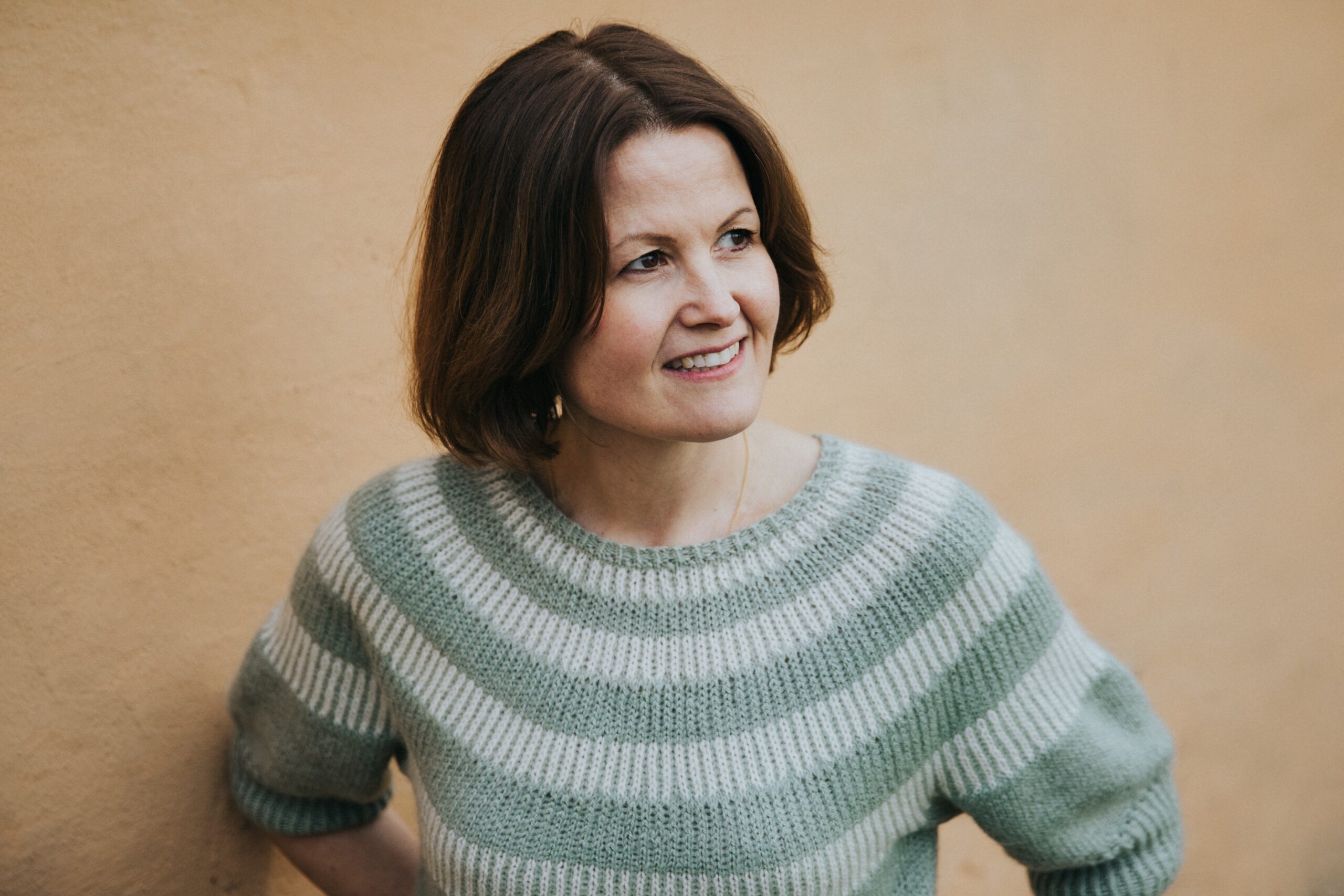 Dr. Marianne Natvik foran en gul murvegg. Ser bort fra kamera, smiler.