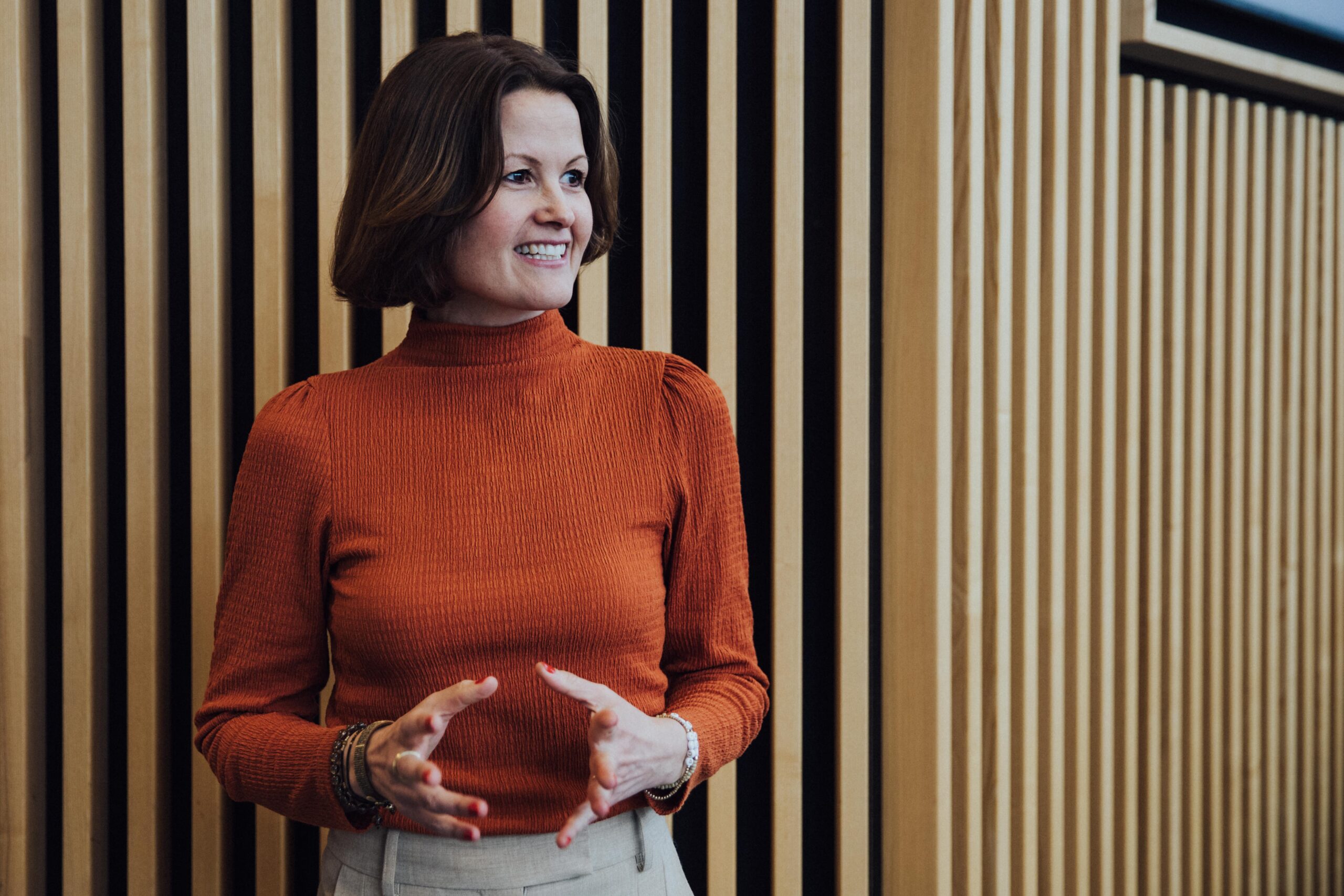 Dr. Marianne Natvik i brunoransje genser, foran en panelvegg. Ser bort fra kamera og smiler.