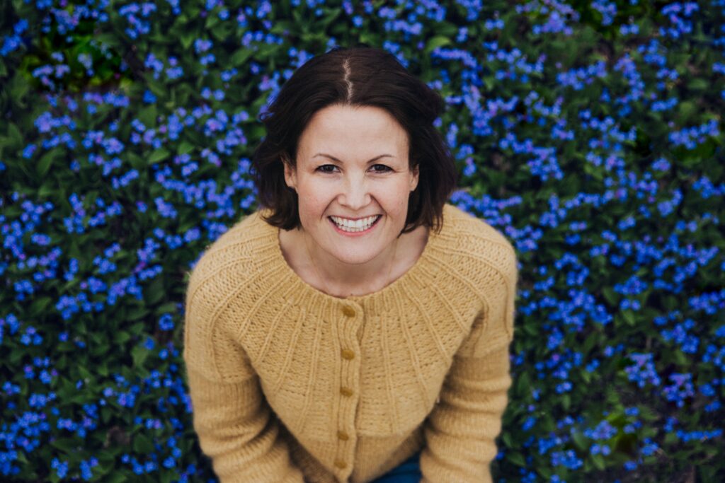 Dr. Marianne Natvik i en blomstereng med mange små blå blomster. Har på deg gul genser, og smiler varmt til kamera.
