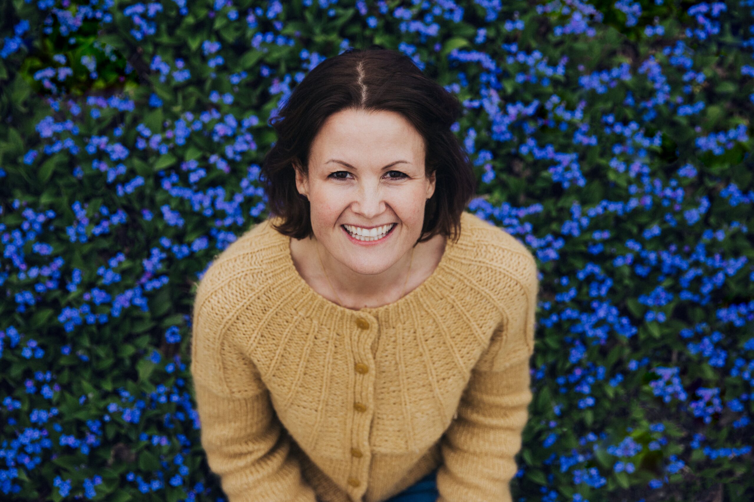 Dr. Marianne Natvik i en blomstereng med mange små blå blomster. Har på deg gul genser, og smiler varmt til kamera.