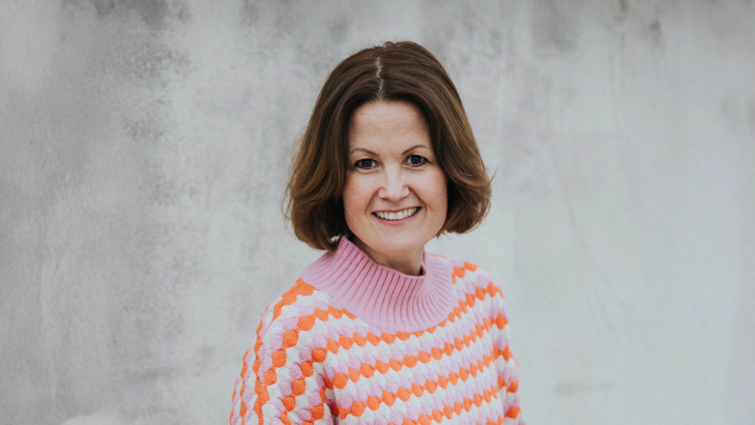 Dr. Marianne Natvik foran en grå murvegg, men fargerik stripete genser. Smiler varmt til kamera.