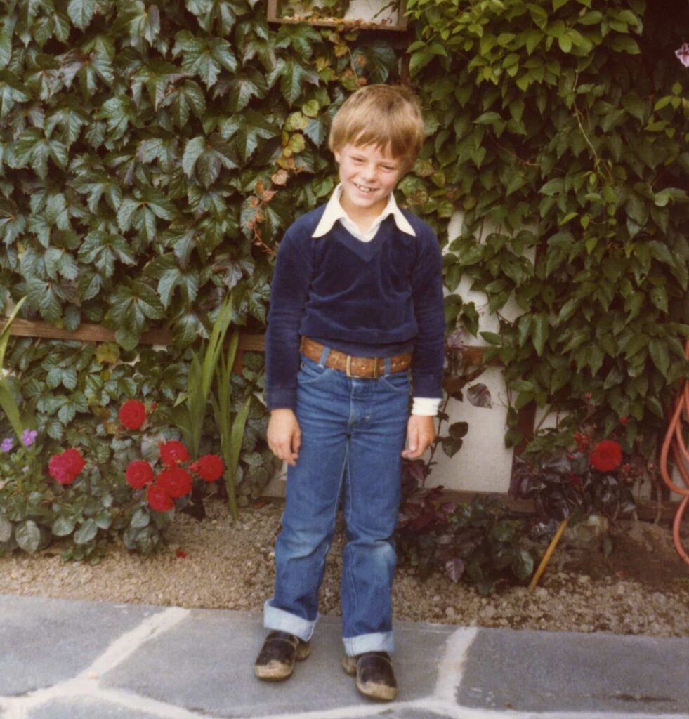 Tore Renberg som barn står i jeans og en blå skjorte foran en vegg dekket av klatreplanter, og smiler. 