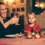 Lyshåret jente sitter ved julepyntet bord, ser inn i kamera mens de voksne tar en skål.