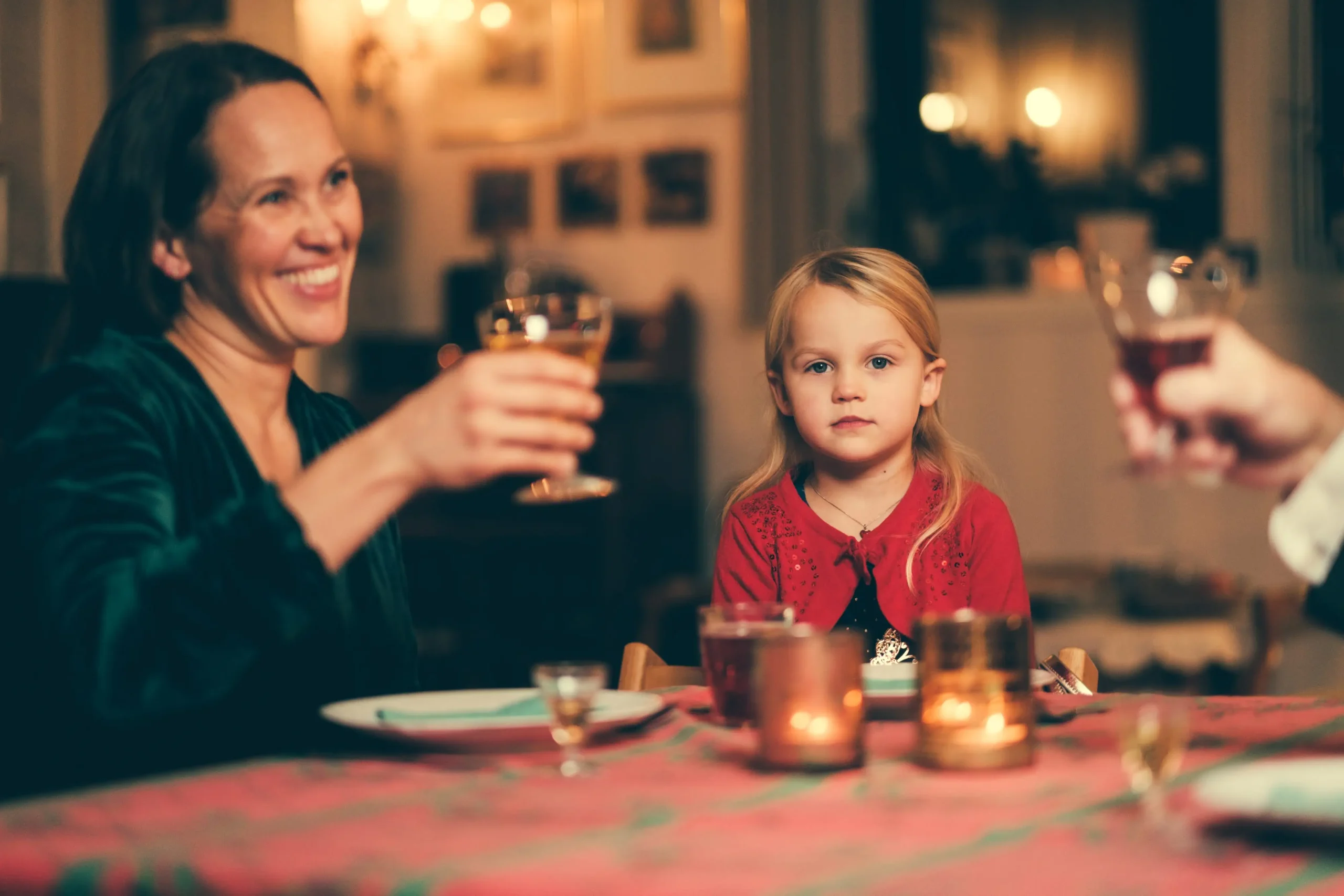 Lyshåret jente sitter ved julepyntet bord, ser inn i kamera mens de voksne tar en skål.