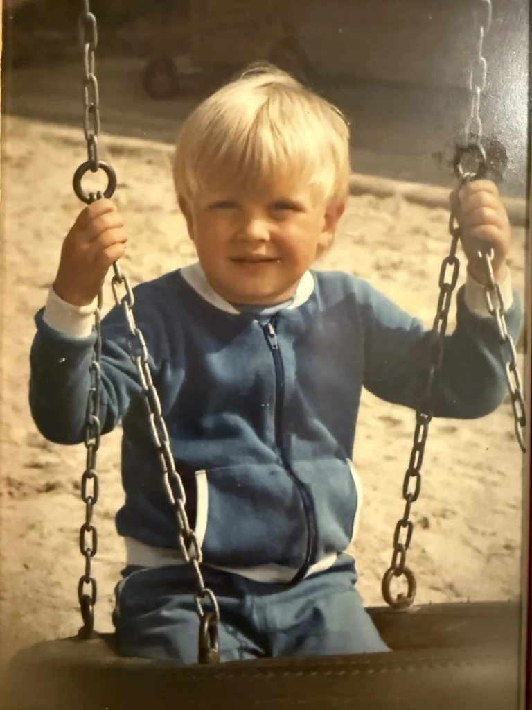 Barnebilde av Ole Erevik på lekeplass. Han sitter på en huske og smiler til den som tar bilde.