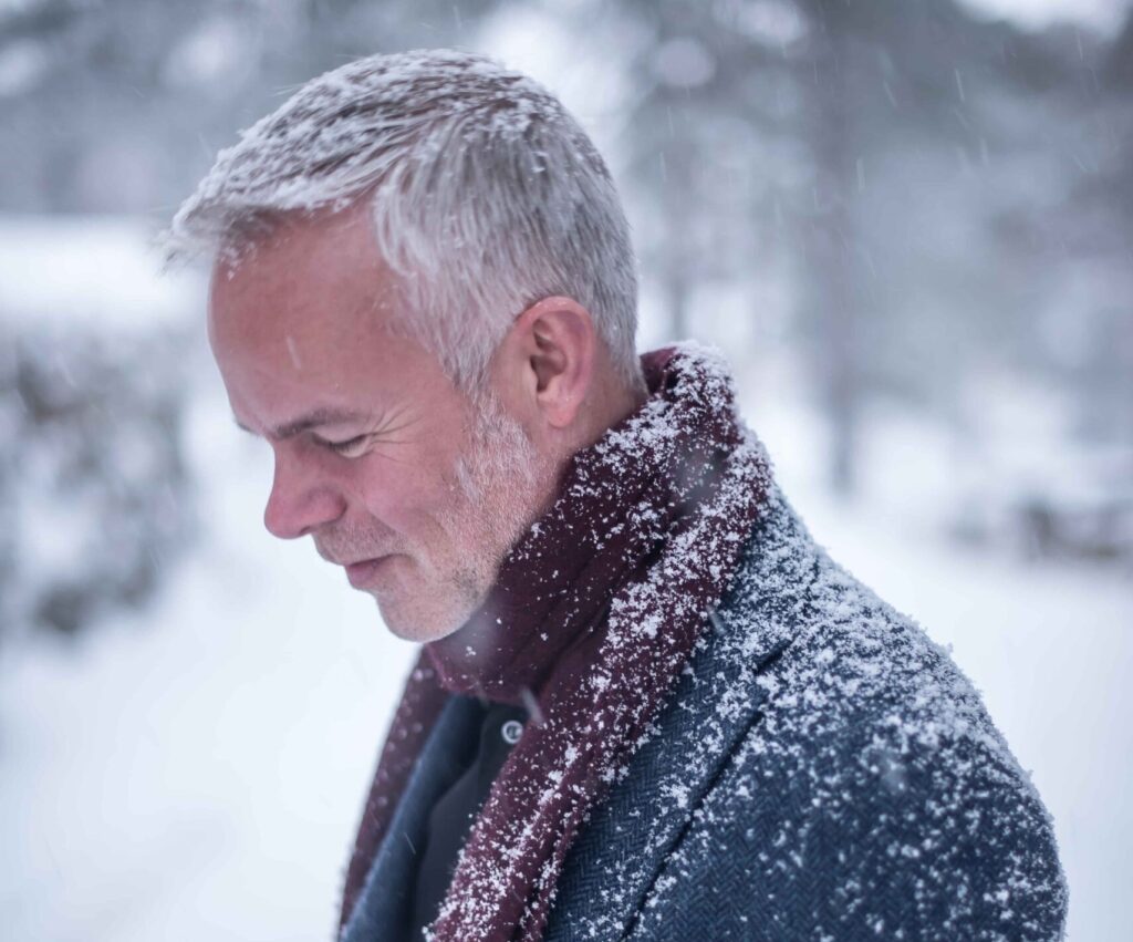 Tore Renberg står i profil, ikledd en dressjakke i ull og et mørkerødt skjerf - i snødekket landskap. 