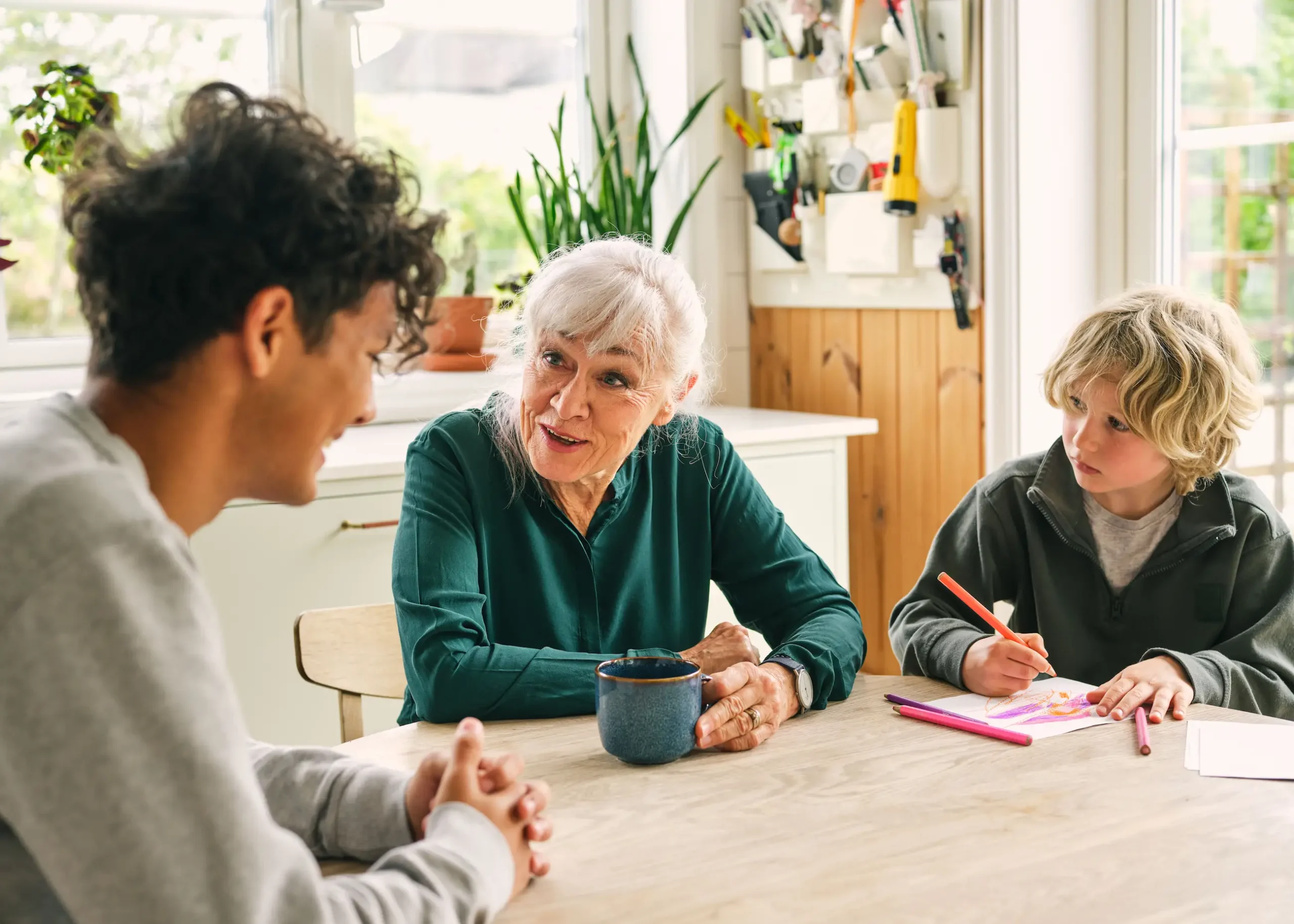 Bestemor og to barnebarn sitter rundt et bord og drikker av en kaffekopp og tegner.