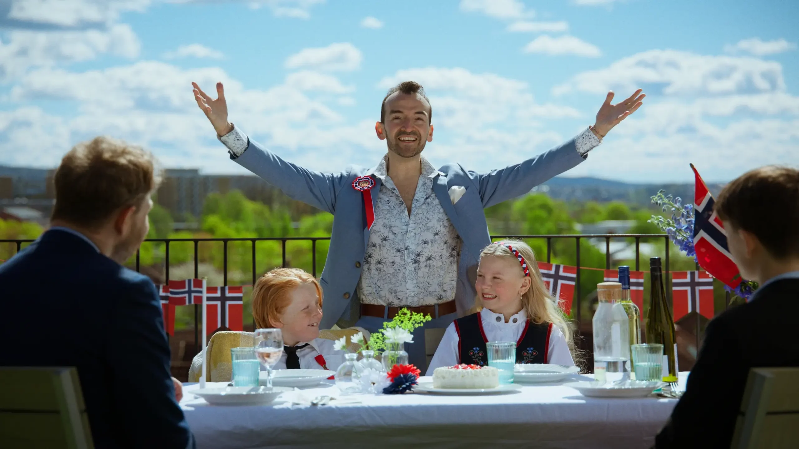 En mann står med armene i været bak et 17. mai-pyntet bord, sammen med barn og voksne som skal feire nasjonaldagen.