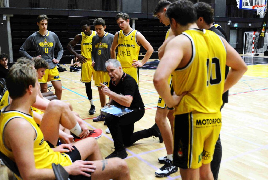 Per Tøisen sitter på huk med basketlaget rundt seg og gir instruksjoner med fingeren pekende. 
