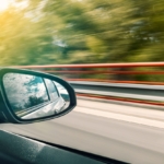 Bilde tatt ut av vinduet på en bil hvor man ser sidespeilet. Utenfor ser man grønne trær, og et svakt sollys - og ut fra bildet kan det virke som om bilen kjører relativt fort.