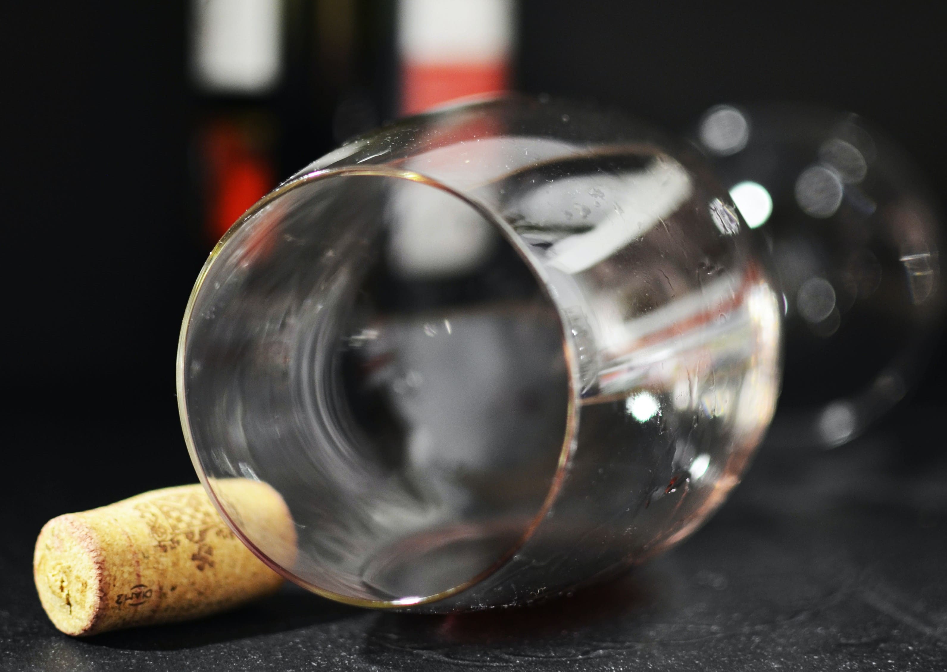 Veltet rødvinsglass med rester, med en vinkork henslengt på siden og en vinflaske i bakgrunnen. Skaper et inntrykk av en fuktig kveld som har kommet litt ut av kontroll.