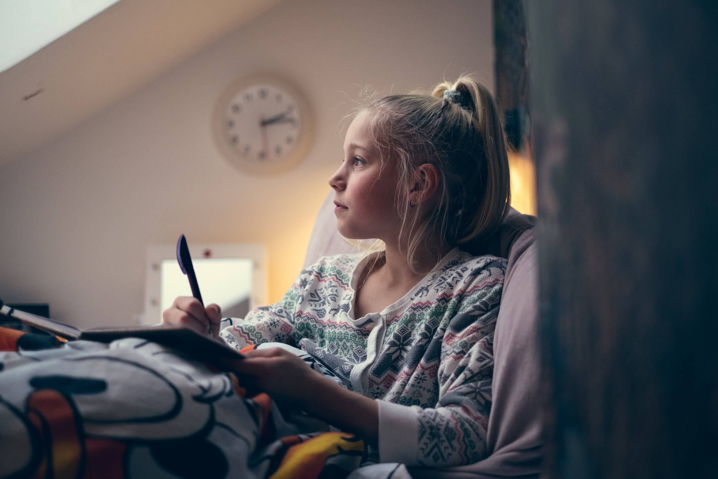 Ung jente sitter i senga si med en dagbok. Hun ser ut i lufta som om hun tenker.