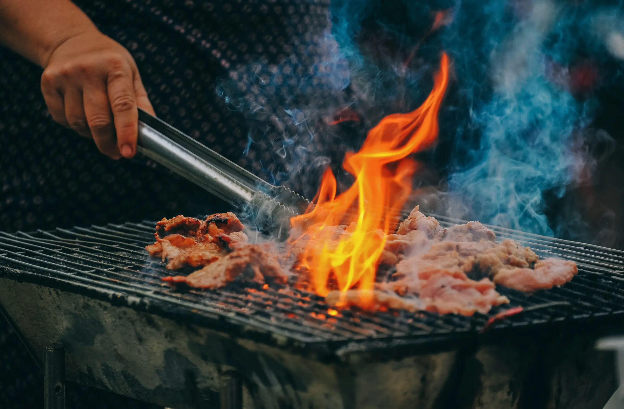 En hånd med en grillklype som tar i kjøtt over en åpen flamme på grillen.