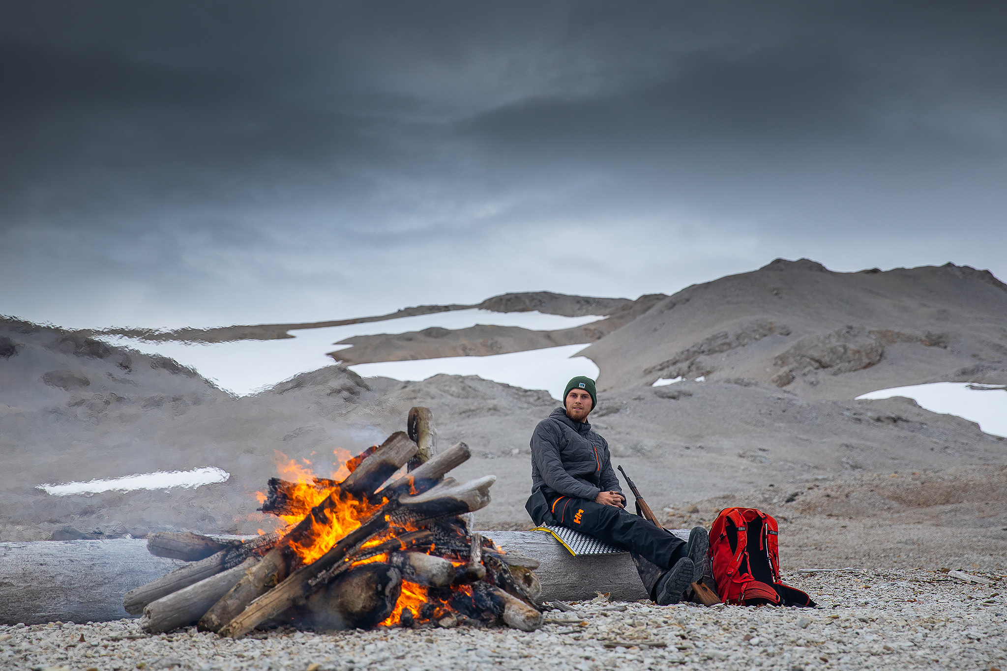Bilde av Mats Grimsæth, vi ser hele han sitte ved et bål og i bakgrunn ser vi fjell med områder med snø.