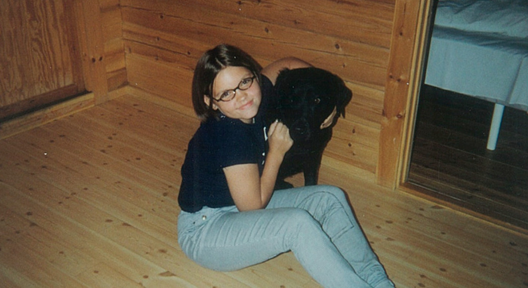 Bilde av Elisabeth som barn. Hun har brunt kort hår og briller, og sitter på gulvet og holder rundt en svart stor hund.