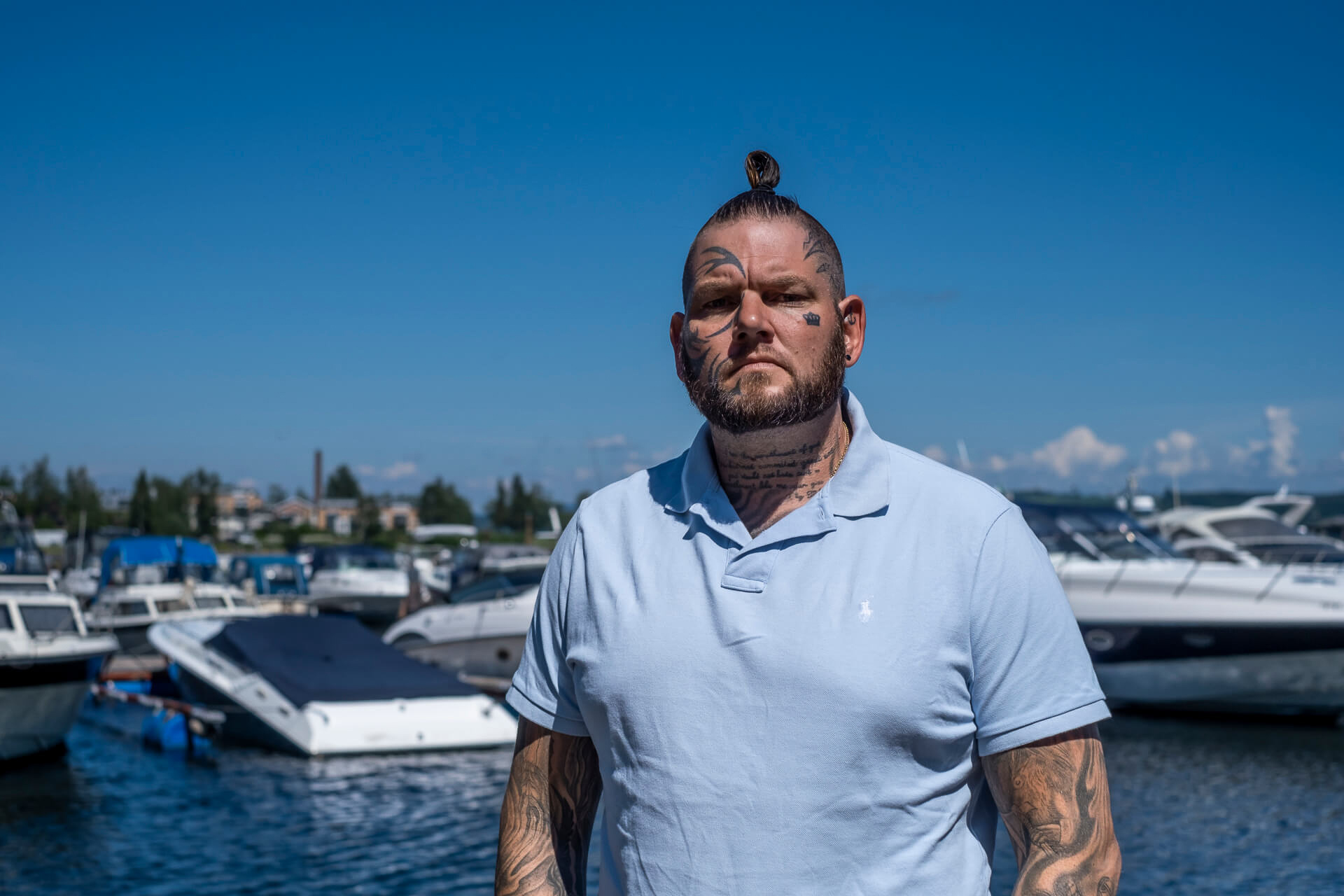 Lars, en ung mann med mange tatoveringer står foran en båthavn med og ser alvorlig inn i kamera