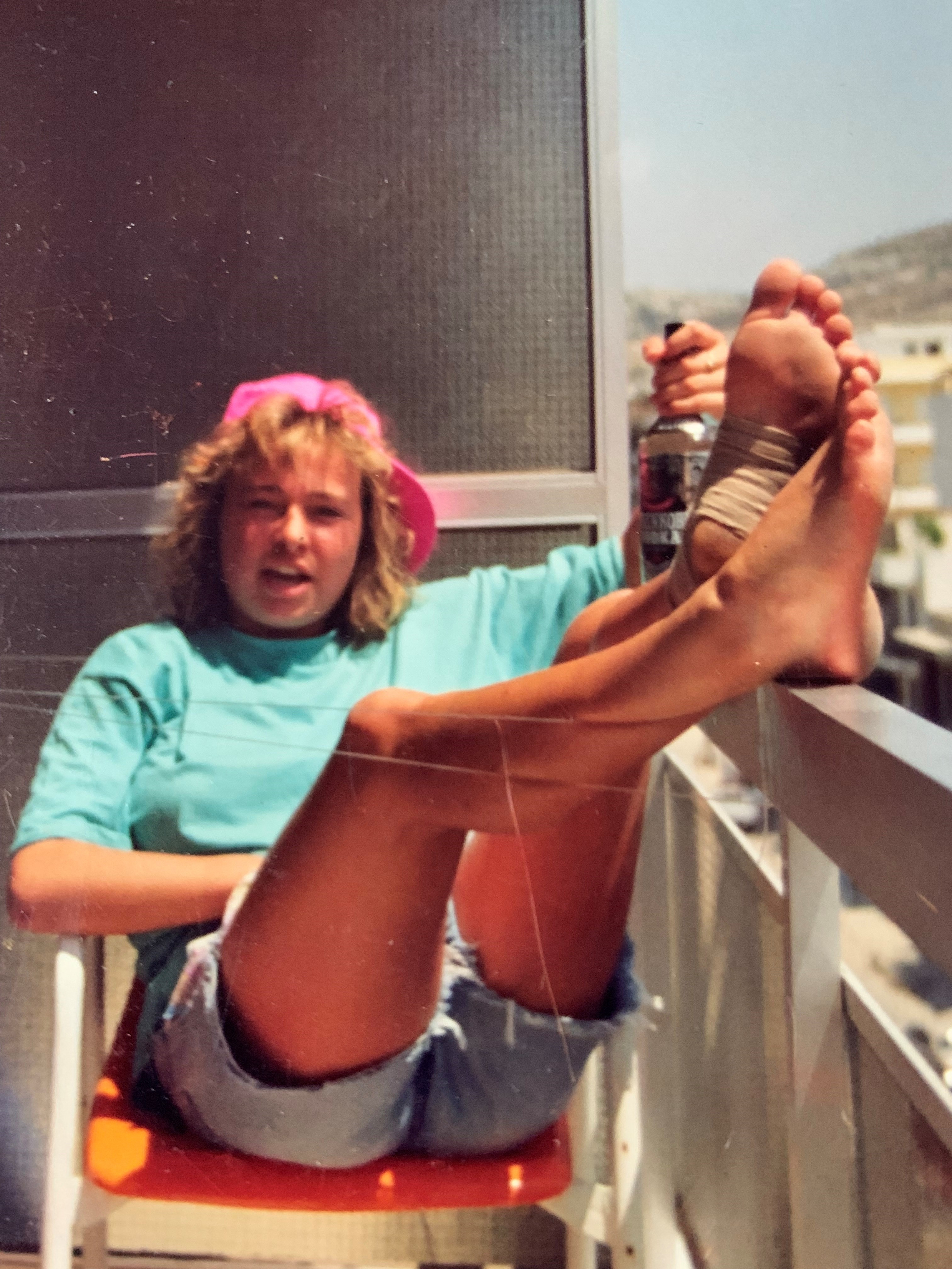 Nina Anett Lea som yngre. En ung dame med shorts, blå t-skjorte og rosa caps bakfrem. Hun har sitter i en stol og har bena over rekkverket på en balkong.