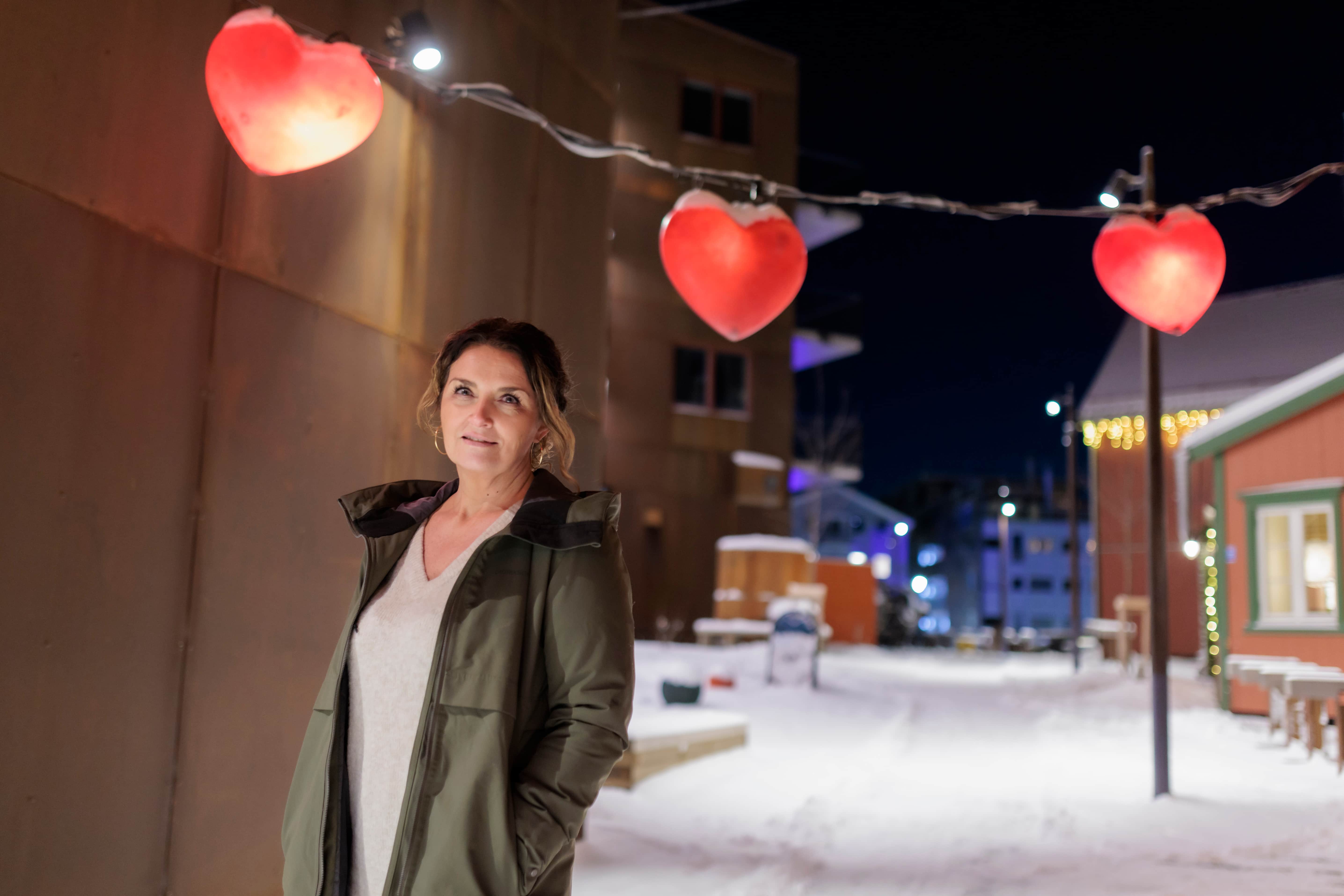 Christel i julegatene i Tromsø. Hun står mellom hus og over henger en lyslenke med større røde hjerter.