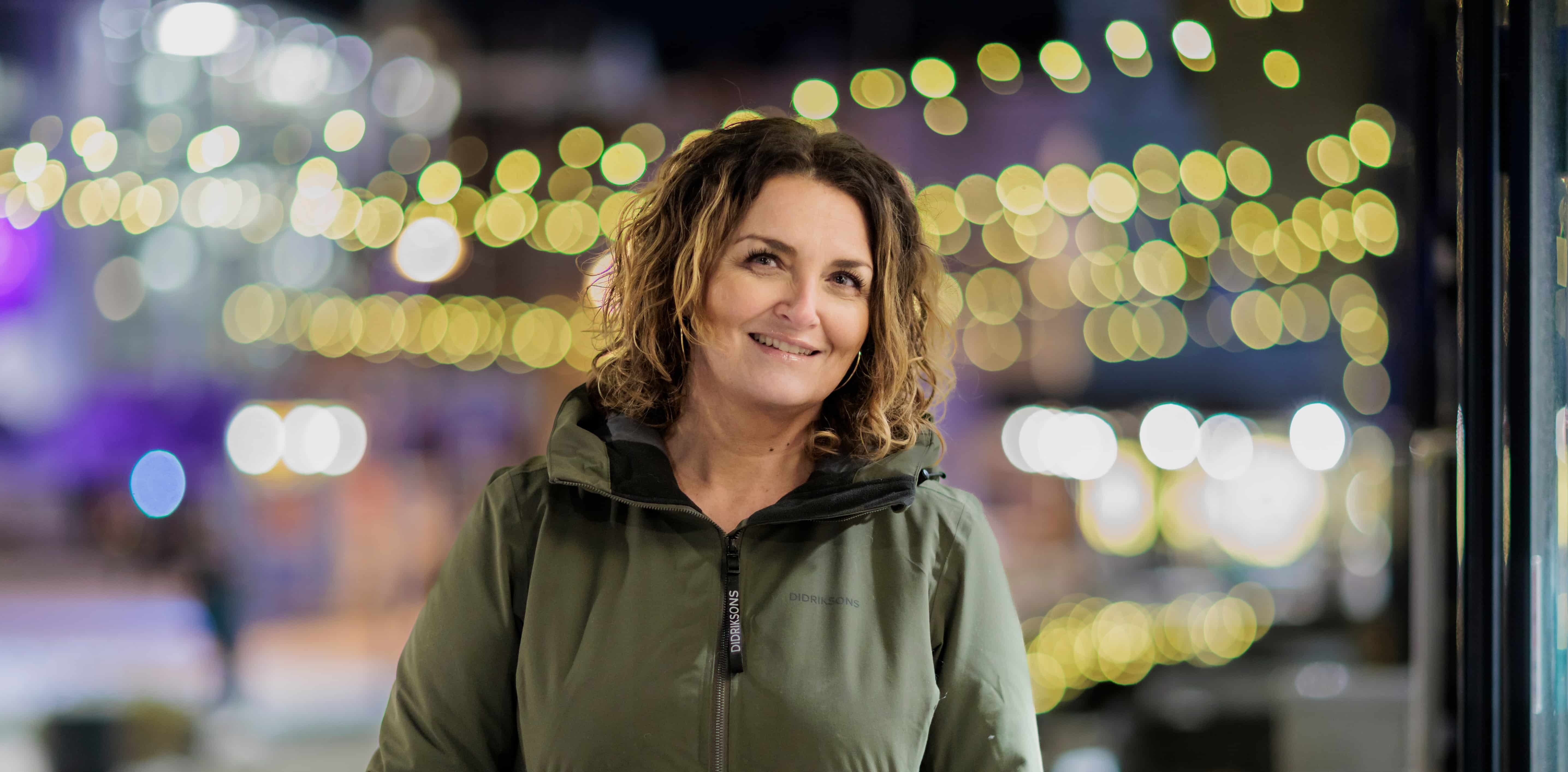 Christel står oppstilt ute i gatene med julelys i bakgrunn, og ser inn i kamera med et lite smil.