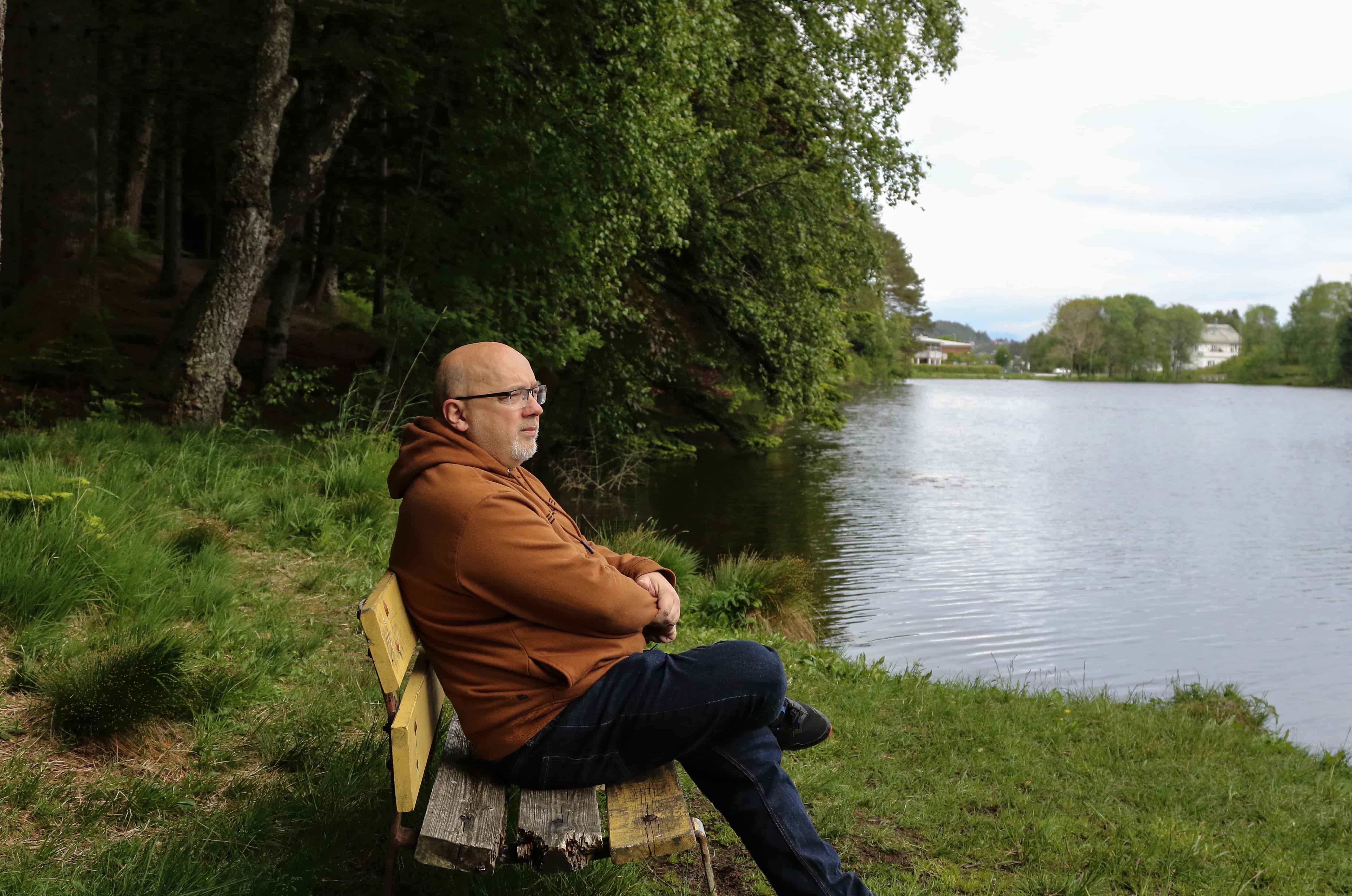 Arne sitter på en trebenk og ser utover et vann. Foto.