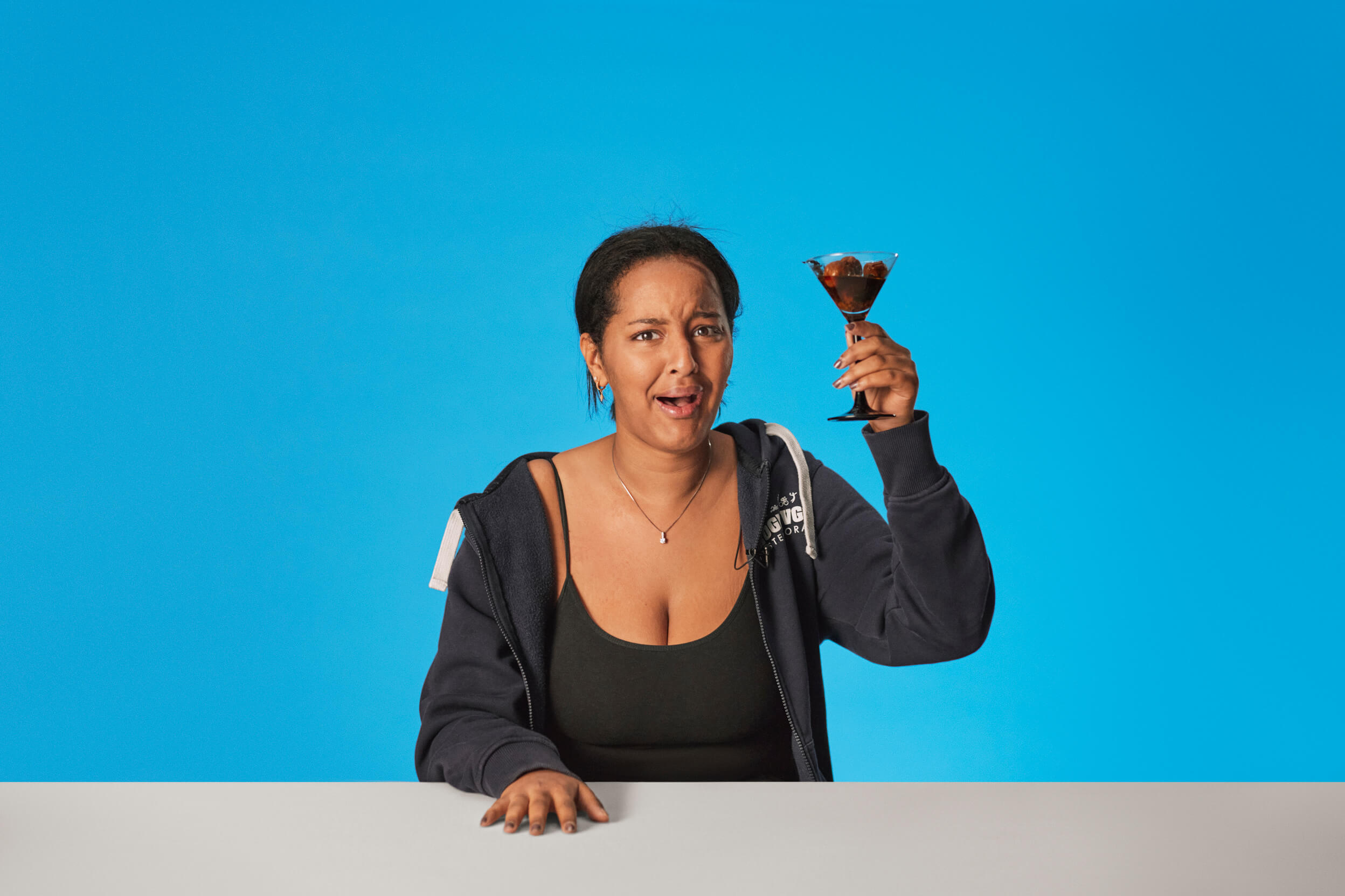Ung kvinne sitter ved bord med blå bakgrunn. Hun holder en drink med ubestemmelig innhold og hun skjærer en grimase med ansiktet.