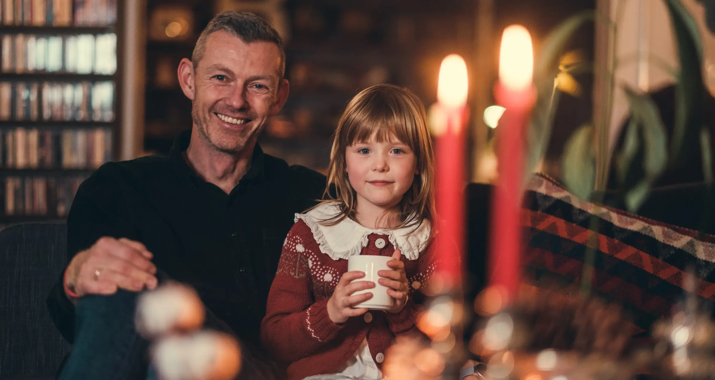 En middeladrende mann og en en jente ser rett inn i kamera. De sitter i et julete rom med stearinlys i forgrunnen som er ute av fokus.