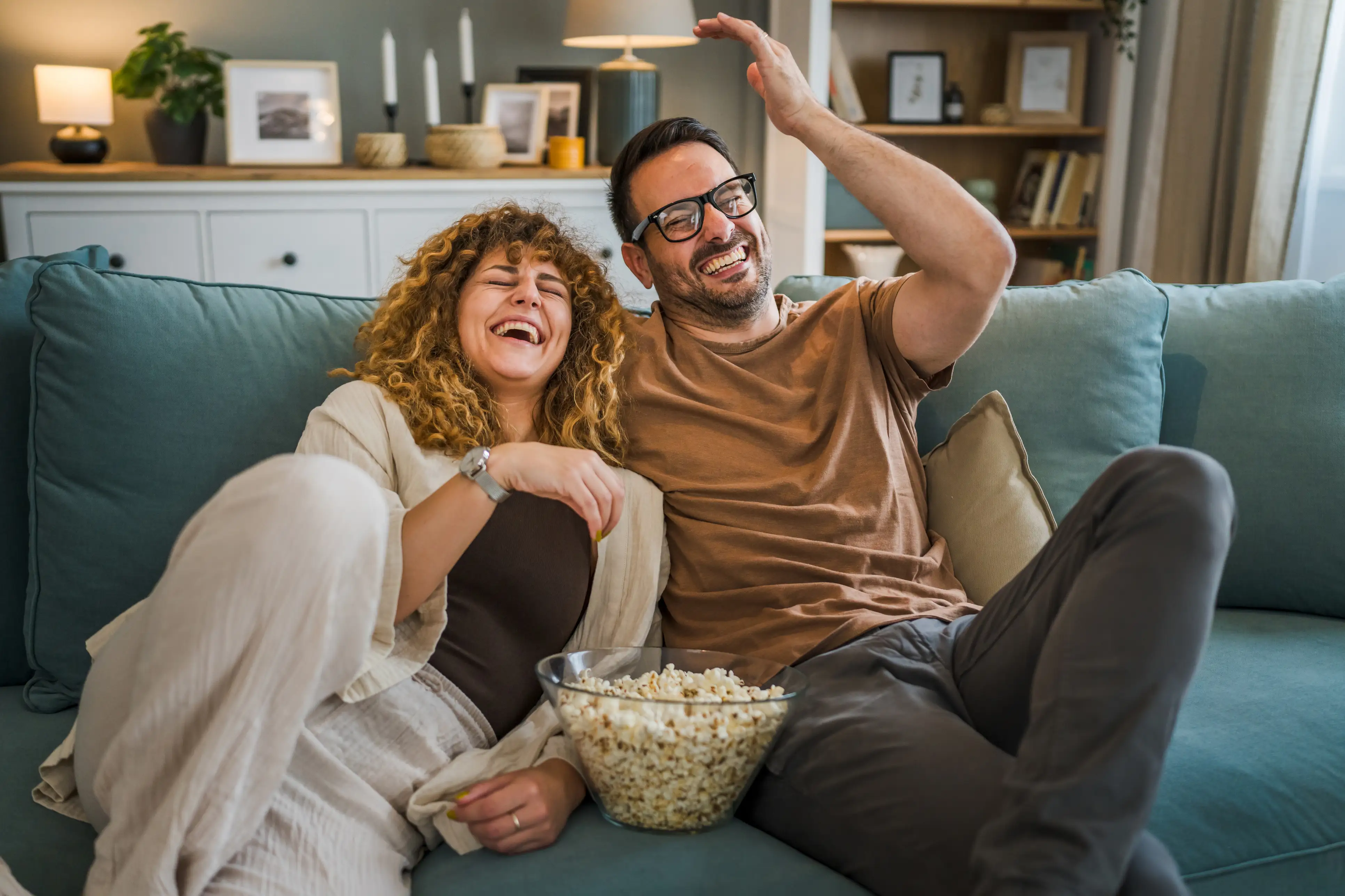Kvinne og mann sitter i sofaen og ler mens de spiser popcorn.