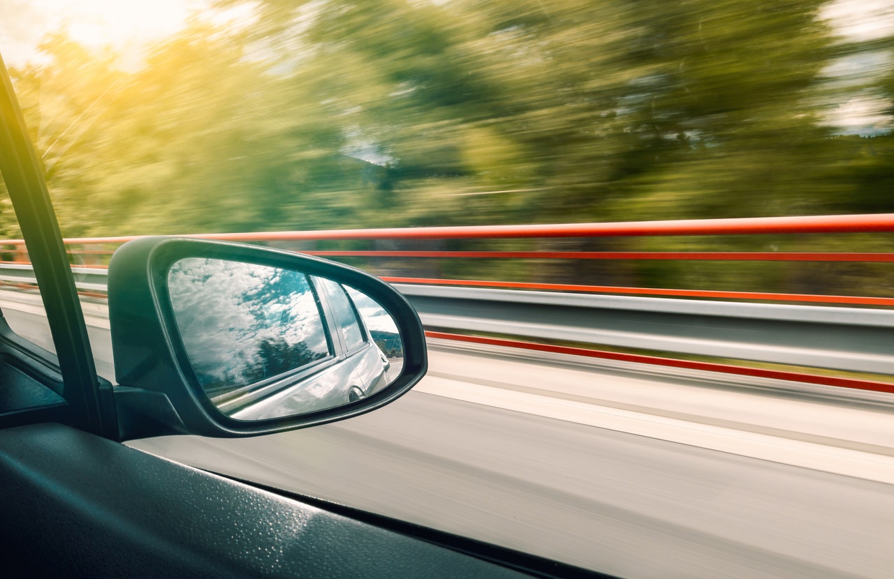 Bilde tatt ut av vinduet på en bil hvor man ser sidespeilet. Utenfor ser man grønne trær, og et svakt sollys - og ut fra bildet kan det virke som om bilen kjører relativt fort.