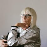 Bilde av en eldre dame med briller og blondt hår og pannelugg. Hun ser mot kamera med et alvorlig blikk.