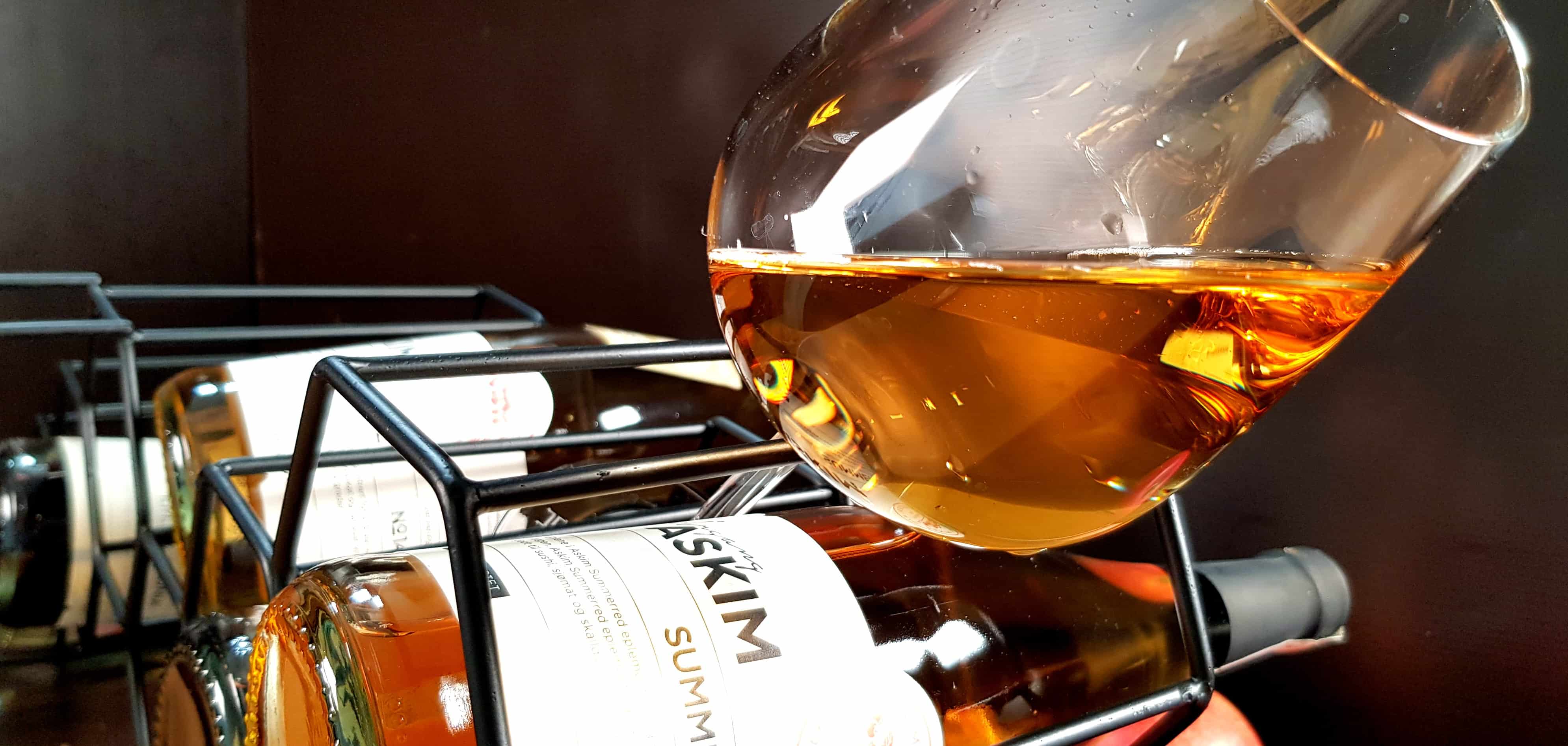 Bilde av et vinglass med cognac-farvet Askim Årgang Summerred. Glasset sitter mellom spilene i et vinstativ, med Askim Årgang-flasker under. Dandert og pent laget for å virke fristende.