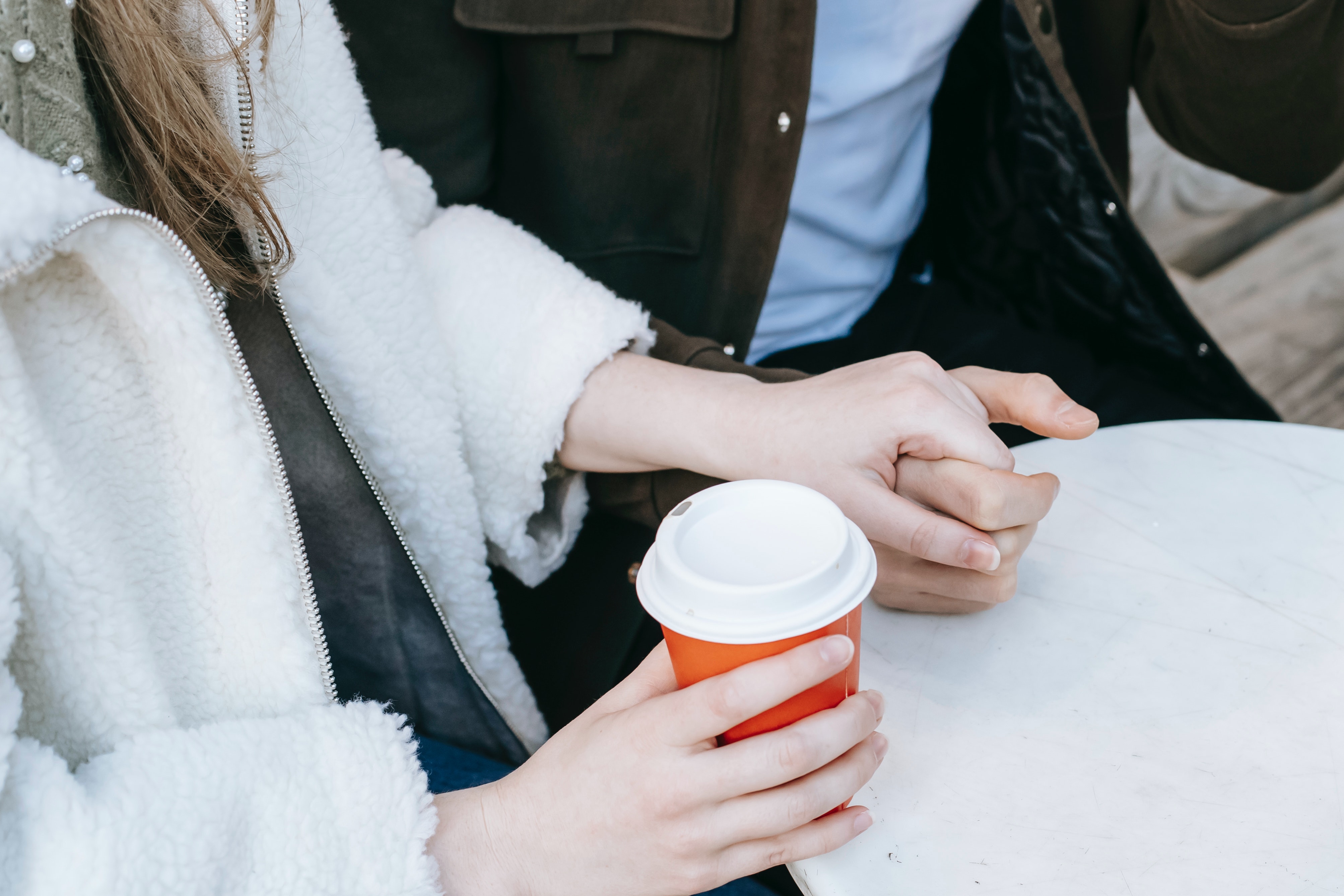 Et par som holder hender. De sitter ved et bord, og damen har venstrehånd rundt en takeaway kaffekopp.