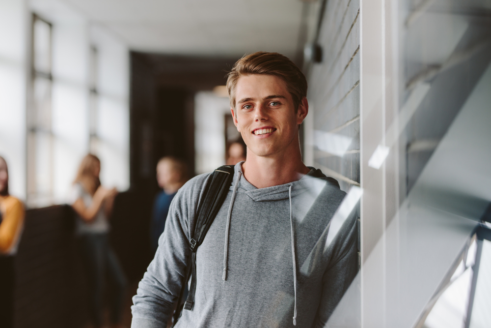 En gutt i ungdomsalder som smiler og ser i kamera mens han lener seg mot veggen i korridoren på skolen med ryggsekken på.