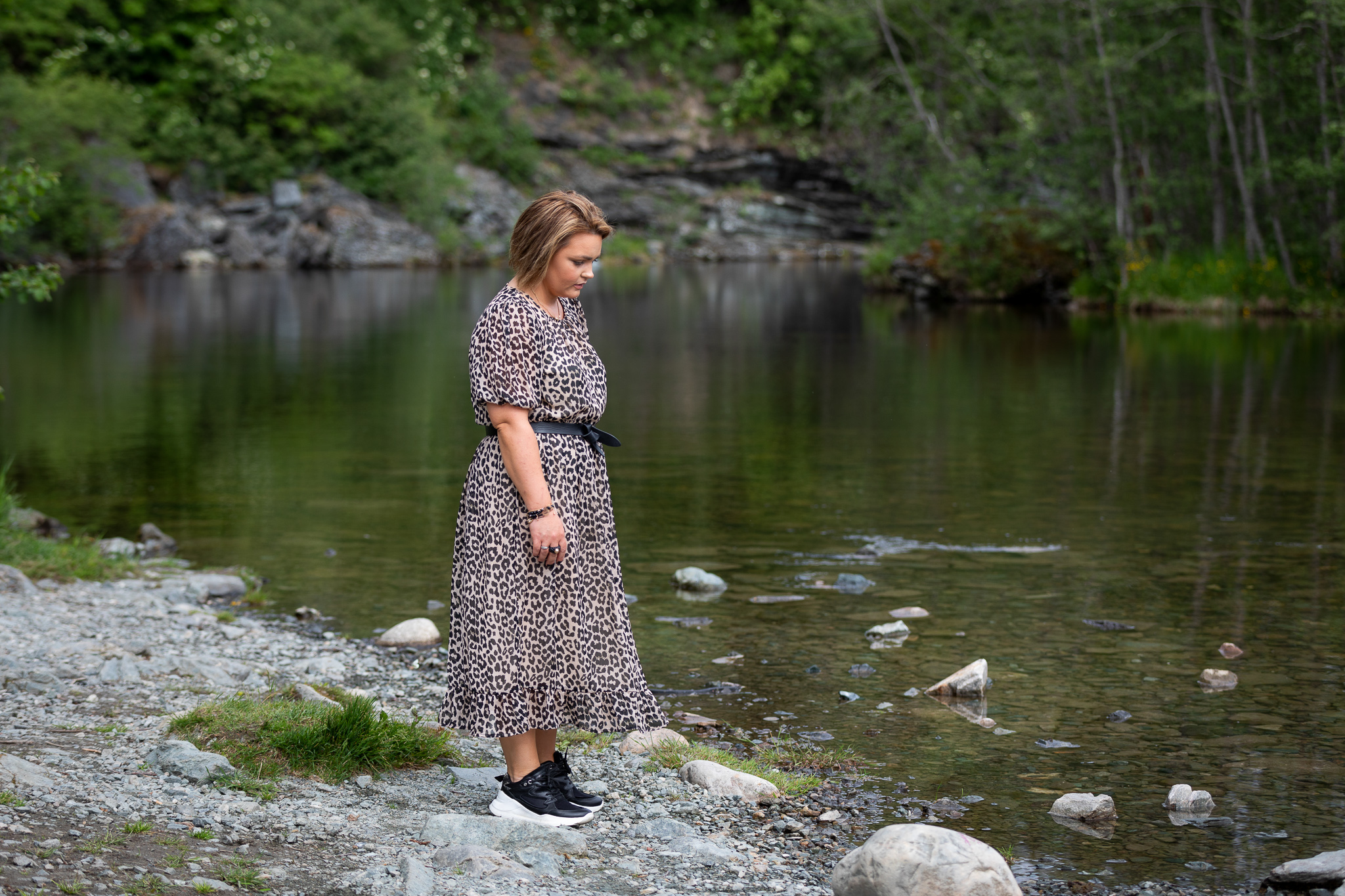 Bilde av en ung dame med kort brunt hår og kjole med leopardmønster (Elisabeth). Hun stå ved en elv og ser ned mot bakken.