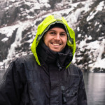 Portrettbilde av eventyrseileren Mats Grimsæth, han smiler bredt inn i kamera ikledd svart regnjakke med gul hette. I bakgrunn ser vi hav.