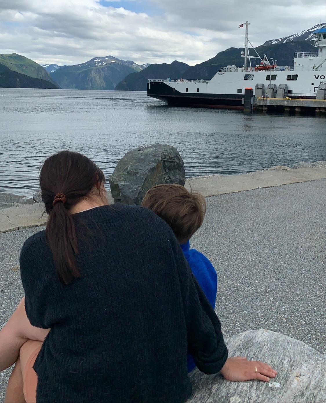 Bilde av en kvinne som sitter med ryggen og ser på sønnen sin som sitter på fanget hennes. I bakgrunn ser vi hav og en større hvit båt.